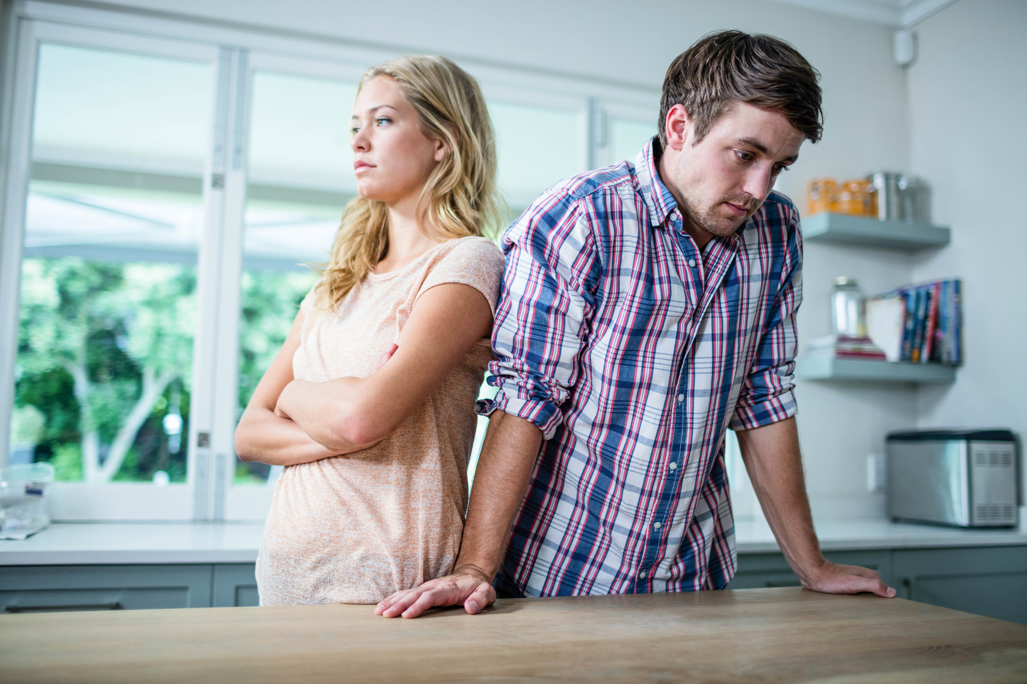 A man and a woman stand in a kitchen. The woman has her arms crossed and looks away, while the man leans on the counter and looks down. The room has modern decor with a window in the background. Both appear to be upset or in disagreement.
