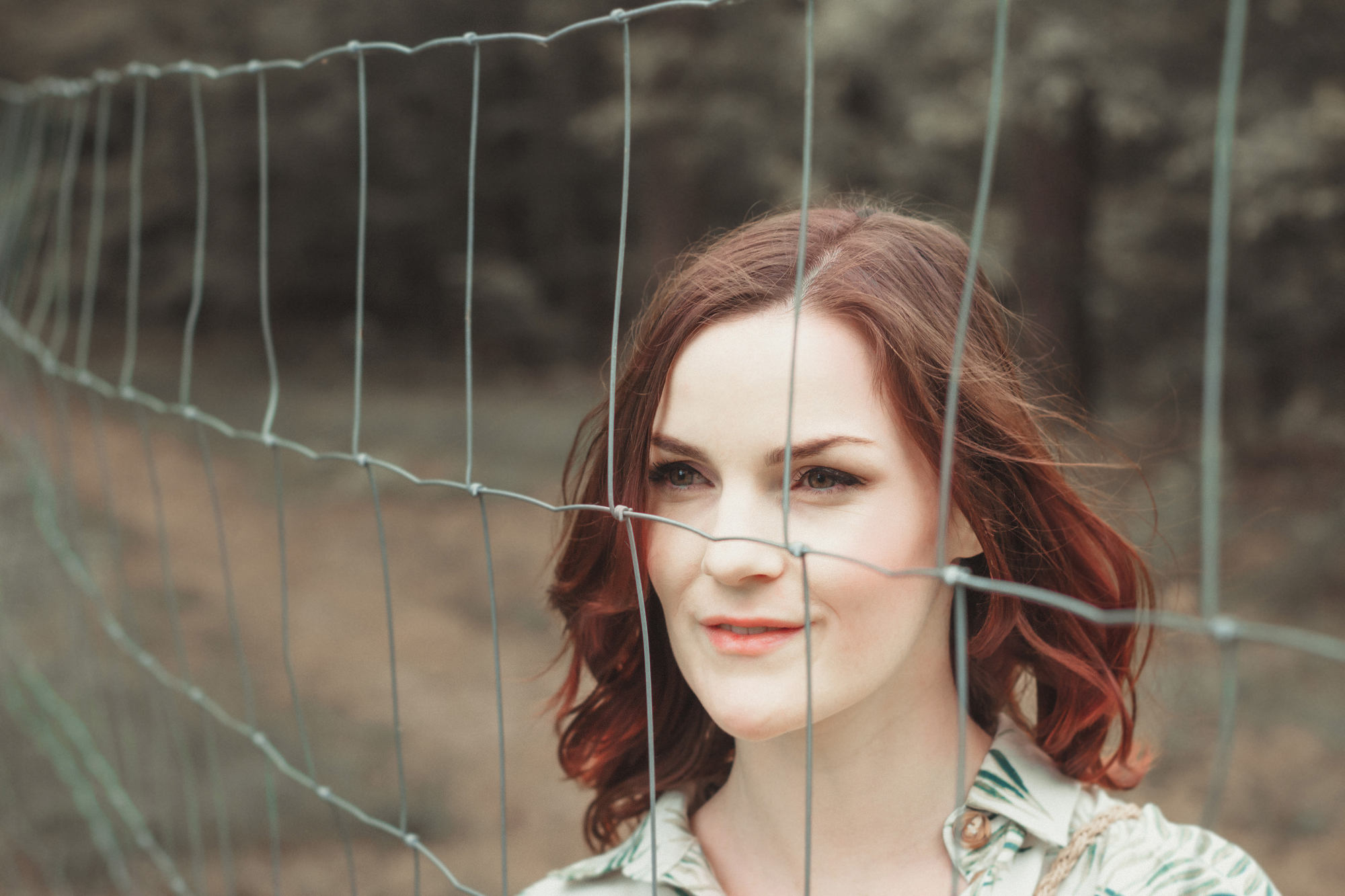 A woman with shoulder-length reddish-brown hair stands behind a wire fence. She wears a light-colored blouse with a subtle pattern and looks slightly to her left with a calm expression. The background is blurry, showing trees and foliage.