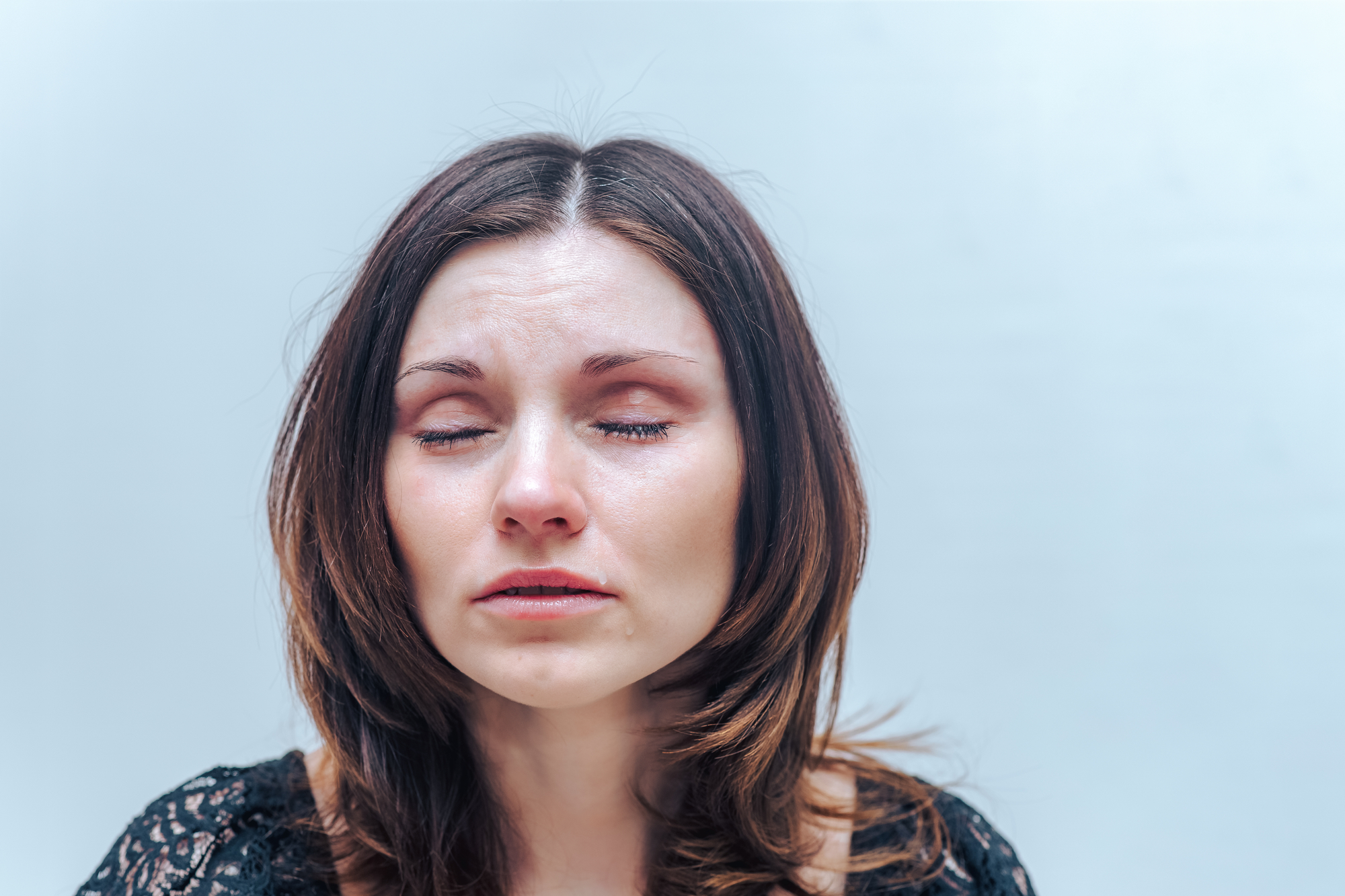 A woman with long brown hair is pictured with her eyes closed, and tears are visible on her face. She appears to be emotional or in distress. She is wearing a black, lacy top, and the background is plain and light-colored.