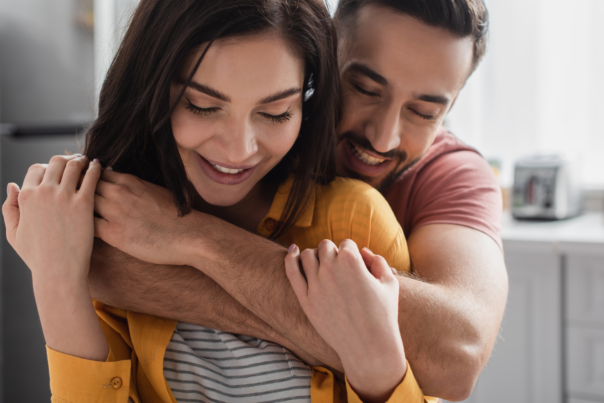 A smiling woman with dark hair, wearing a yellow shirt over a striped top, is being affectionately hugged from behind by a man with short hair and a beard, wearing a pink shirt. They appear happy and are in a bright, modern kitchen.