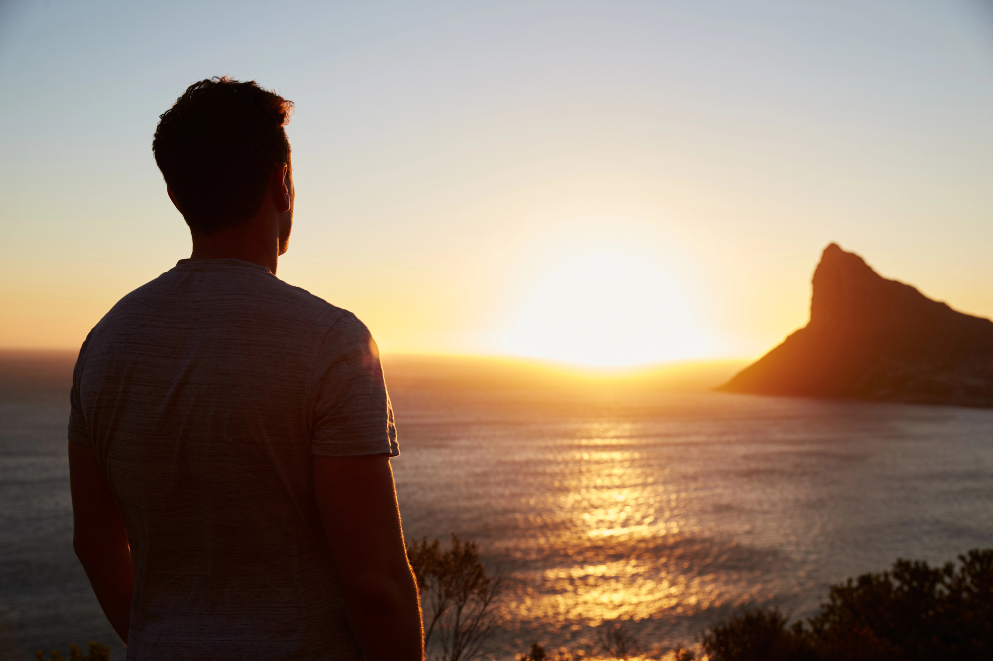 A man in a gray t-shirt stands on a hilltop, gazing at a serene sunset over the ocean. The sun is low on the horizon, casting a warm glow across the water and silhouetting a rugged mountain in the background.