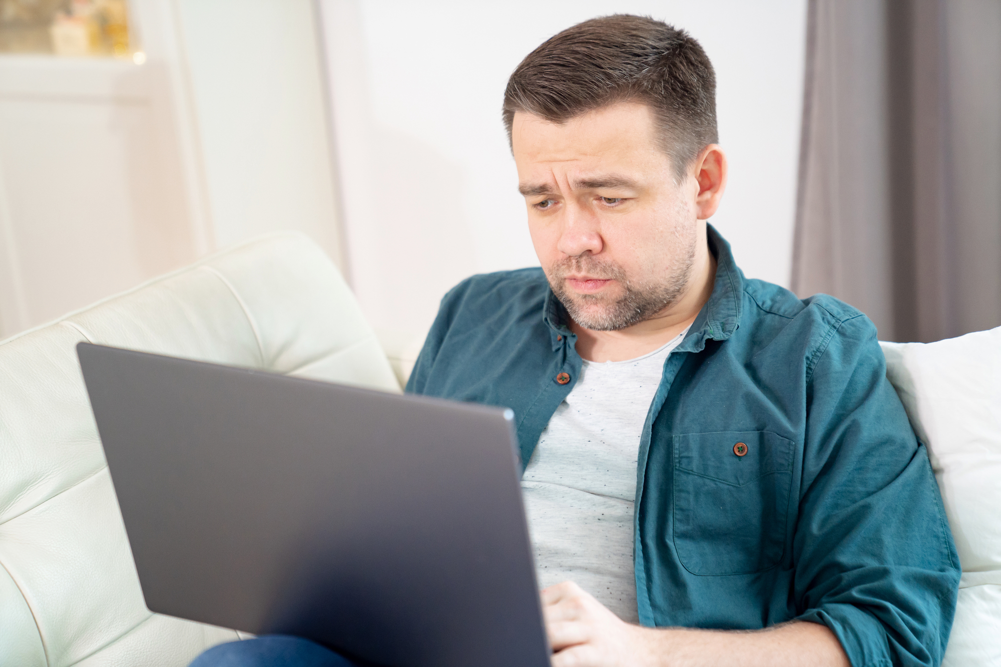 A man with short dark hair and a beard sits on a white couch, concentrating on his laptop screen. He is wearing a teal button-down shirt over a grey t-shirt. The background includes a window with sheer curtains.