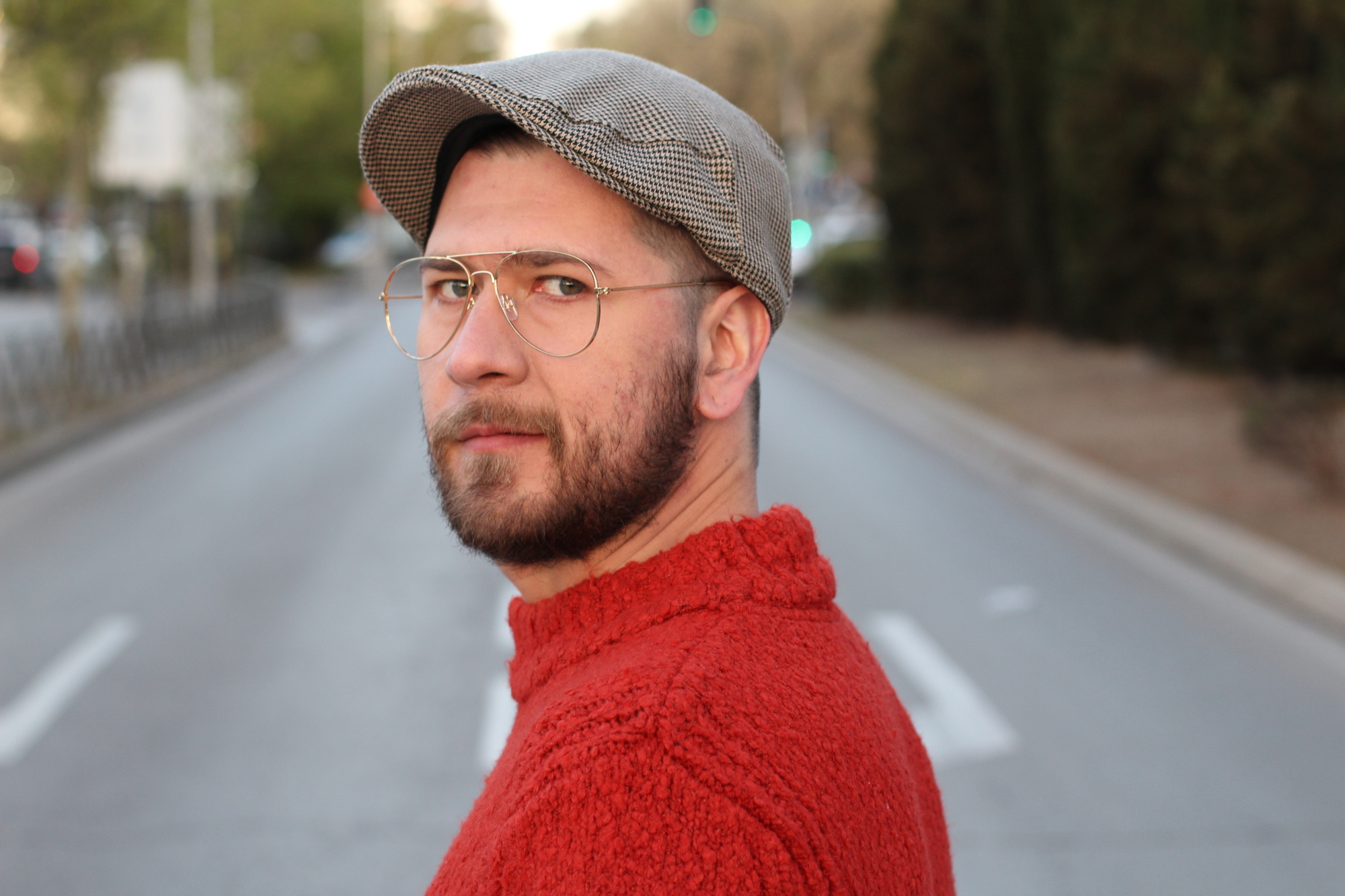 A man with a beard and glasses wearing a red sweater and a gray flat cap looks over his shoulder while standing on an empty road lined with trees. The background is slightly blurred, giving emphasis to his facial expression.