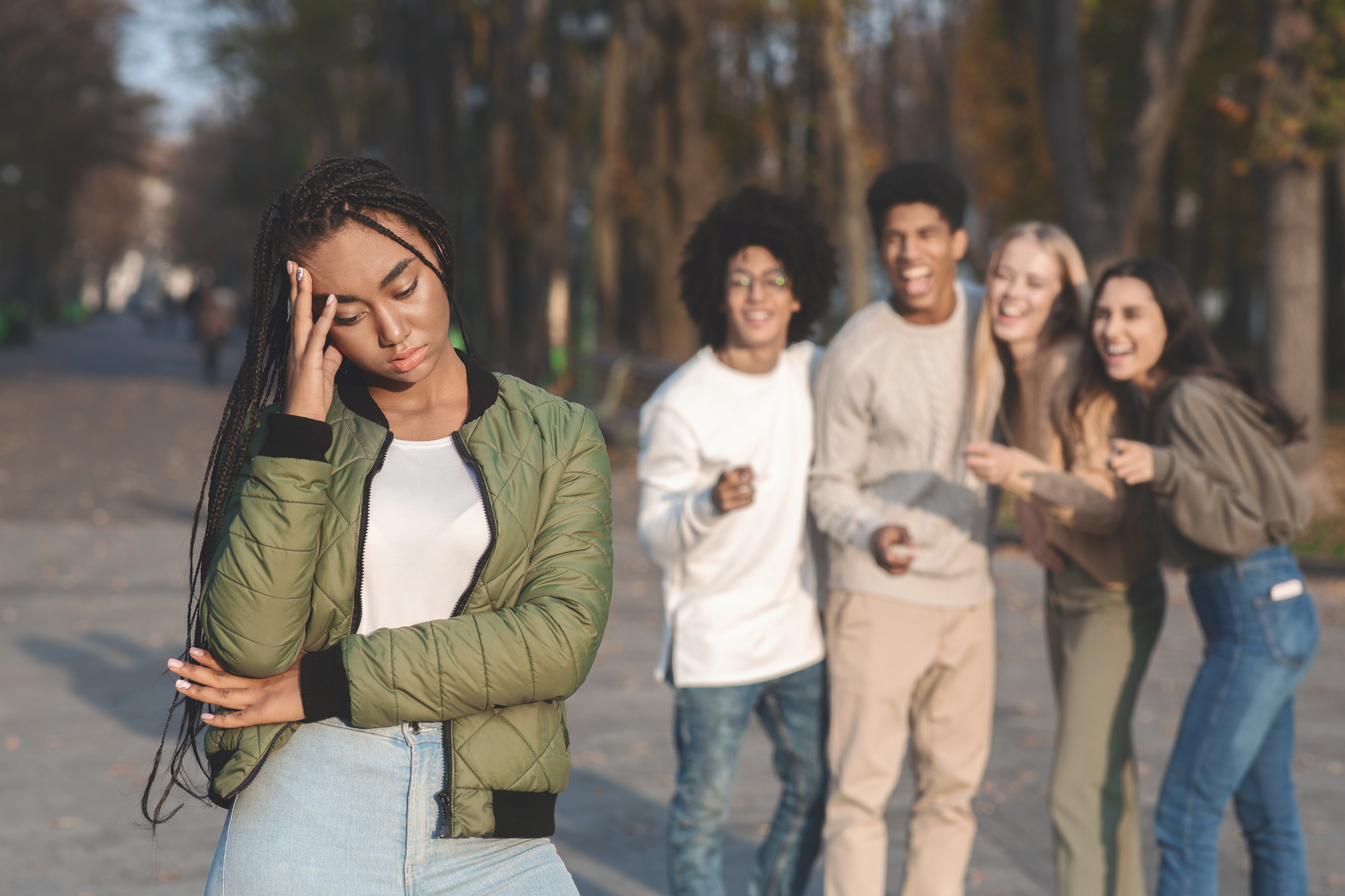 A young woman stands with her eyes closed, looking distressed, and her hand on her forehead. She is in focus and in the foreground. In the background, four young people are pointing at her and laughing. They are outside in a park or natural setting.