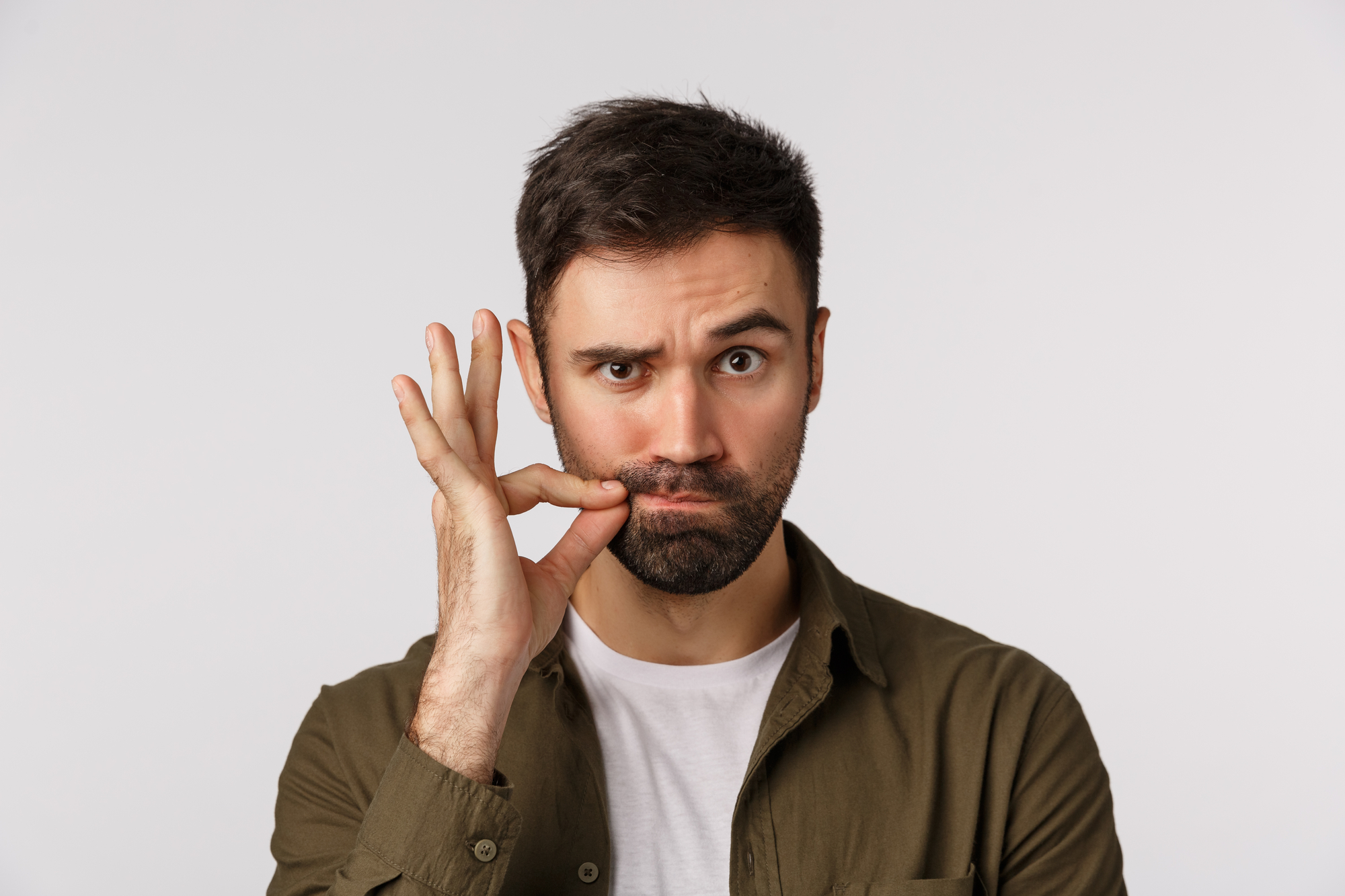 A man with dark hair and a beard stands against a plain background. He is wearing an olive green shirt over a white t-shirt. He has a puzzled expression and is making an "OK" sign with his right hand while pressing his lips together with that hand's thumb and forefinger.
