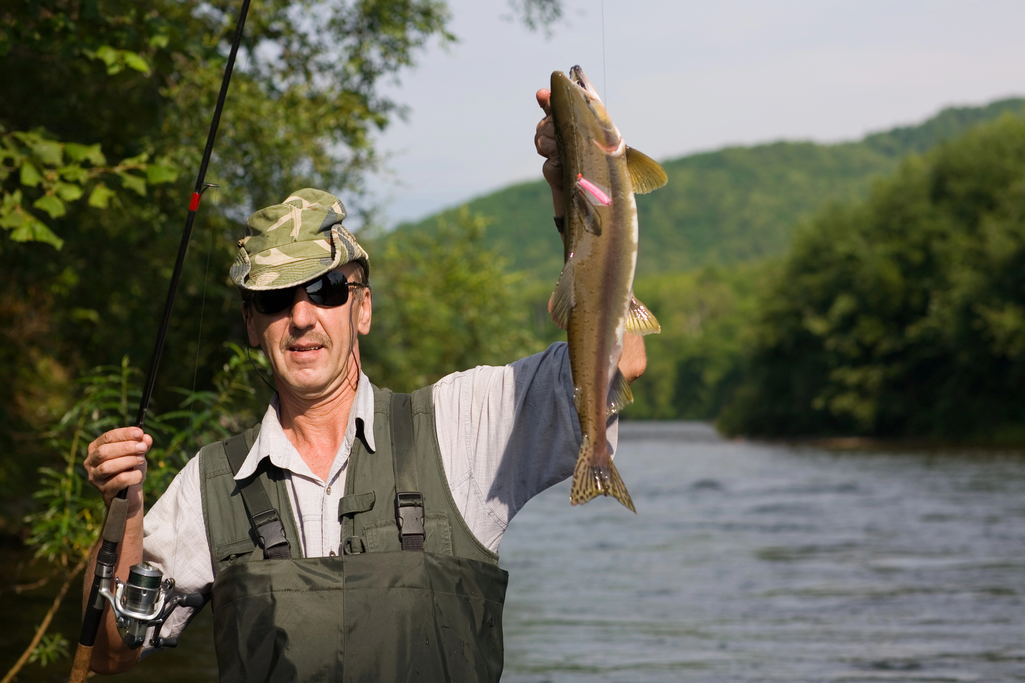A man wearing a camouflage hat and sunglasses holds up a fish he caught while fishing in a river. Dressed in a grey shirt and green waders, he stands among a backdrop of lush trees and a flowing river with hills in the distance.