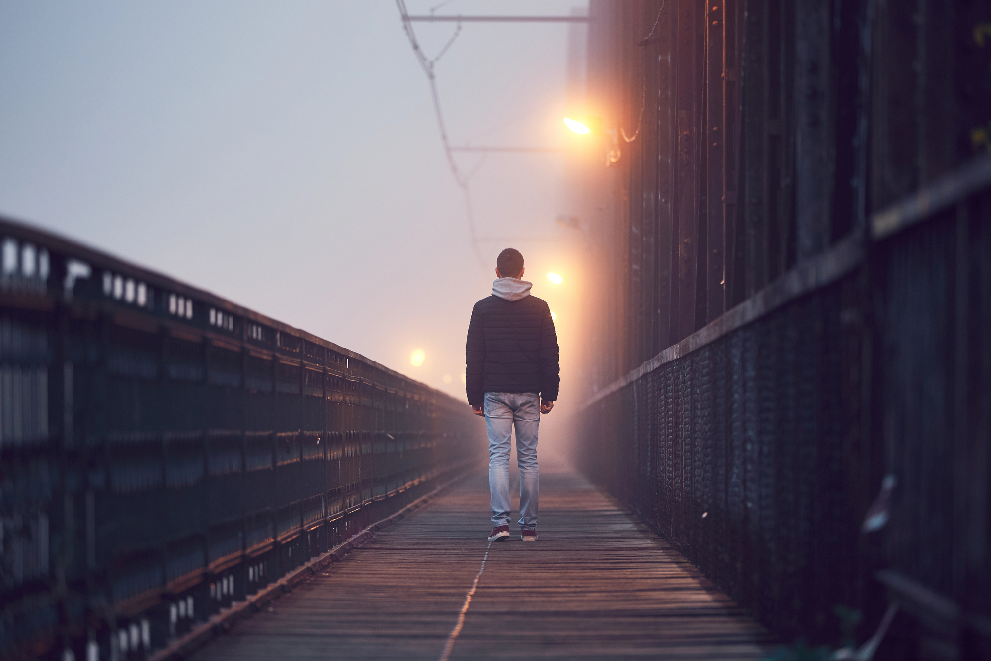 A person in a black jacket and blue jeans walks alone on a foggy, dimly lit bridge. Streetlights cast a warm glow, illuminating the path. The atmosphere is moody and serene, with the fog obscuring the background further down the bridge.
