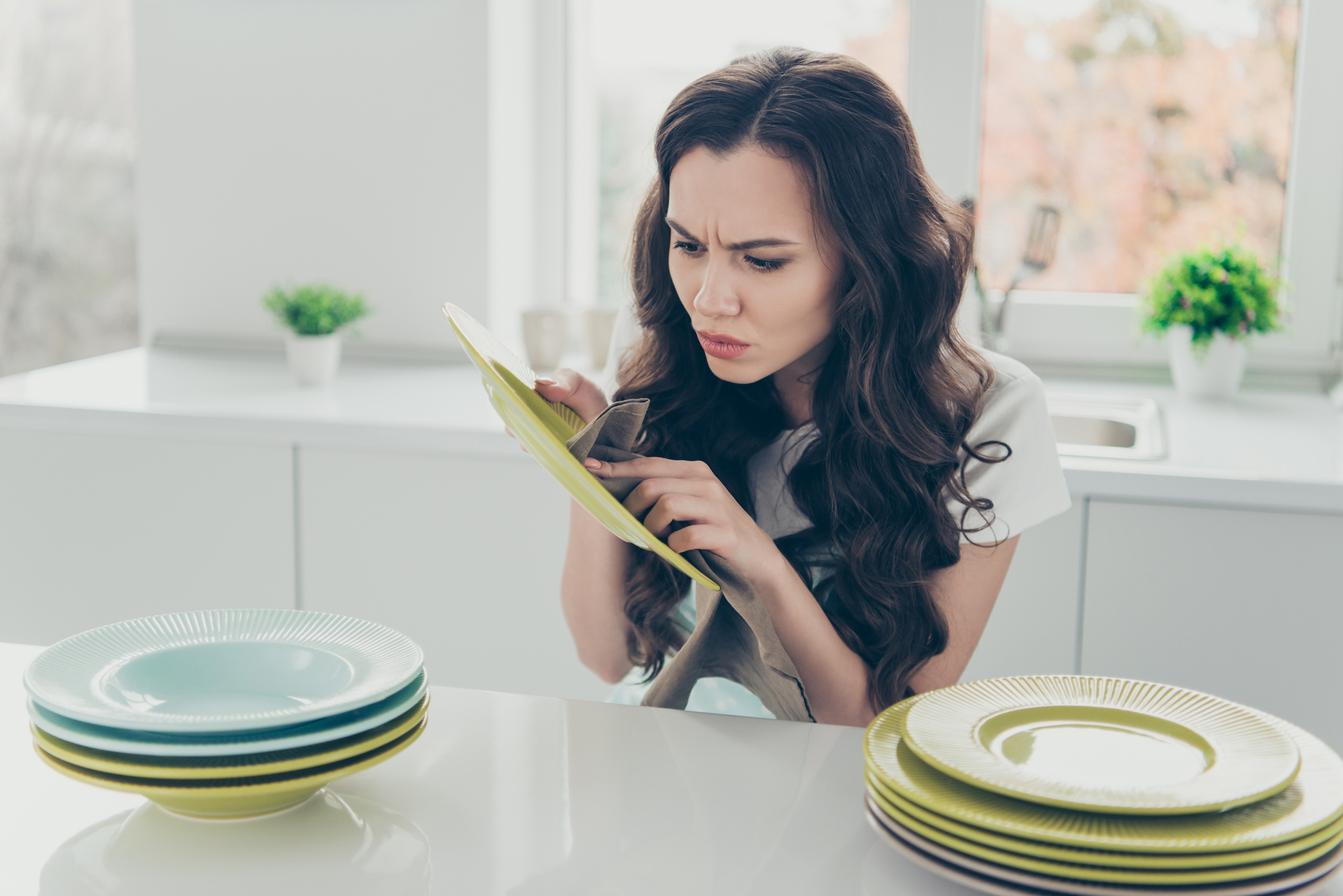 A woman with long, wavy hair examines a yellow plate closely with a frowning expression. She is standing in a bright kitchen with stacks of plates in front of her and potted plants on the windowsill in the background.