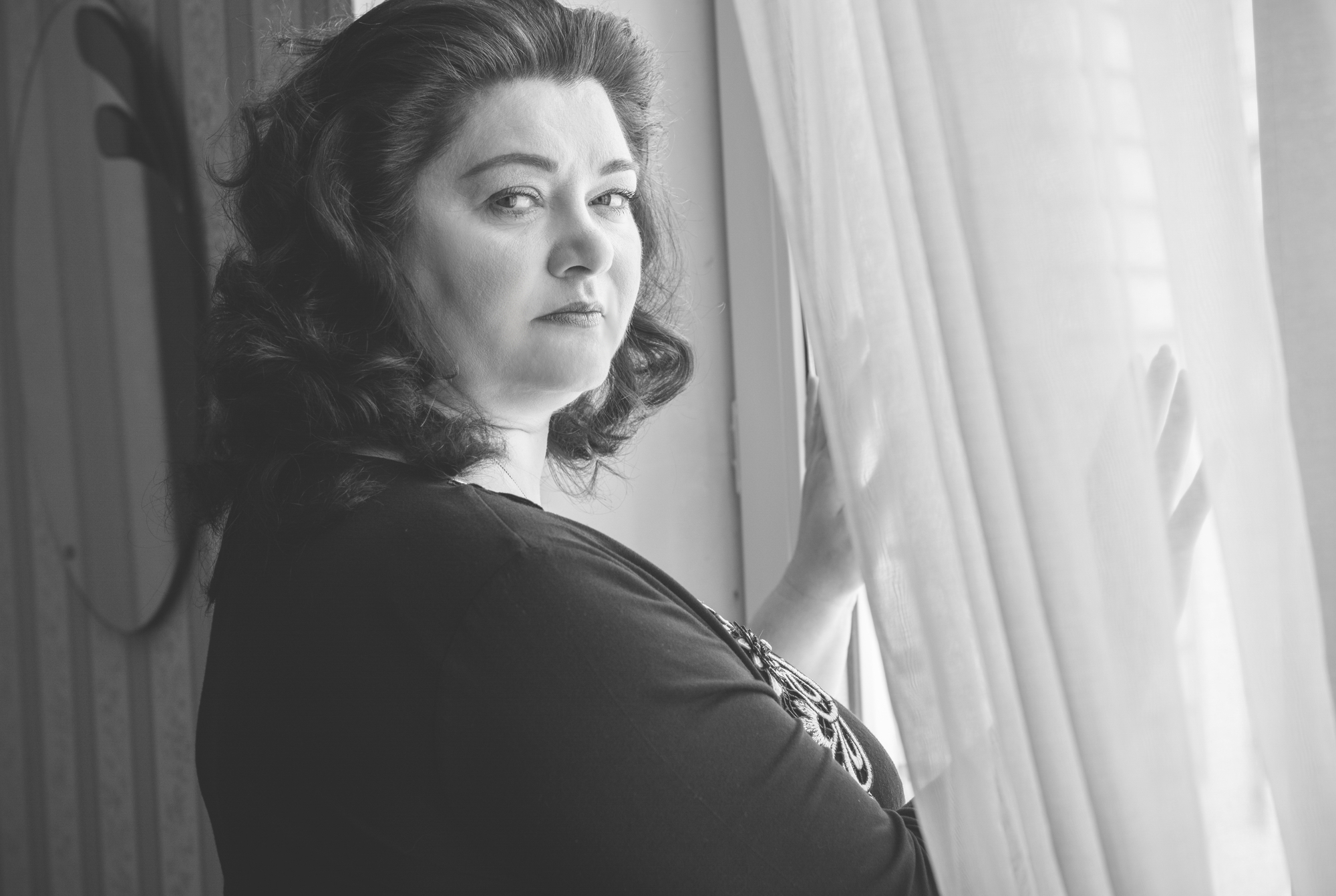 Black and white photo of a woman with wavy, dark hair and a serious expression. She is standing near a window, holding a curtain and looking towards the camera with a contemplative gaze. She is wearing a dark, long-sleeved top with a patterned design.