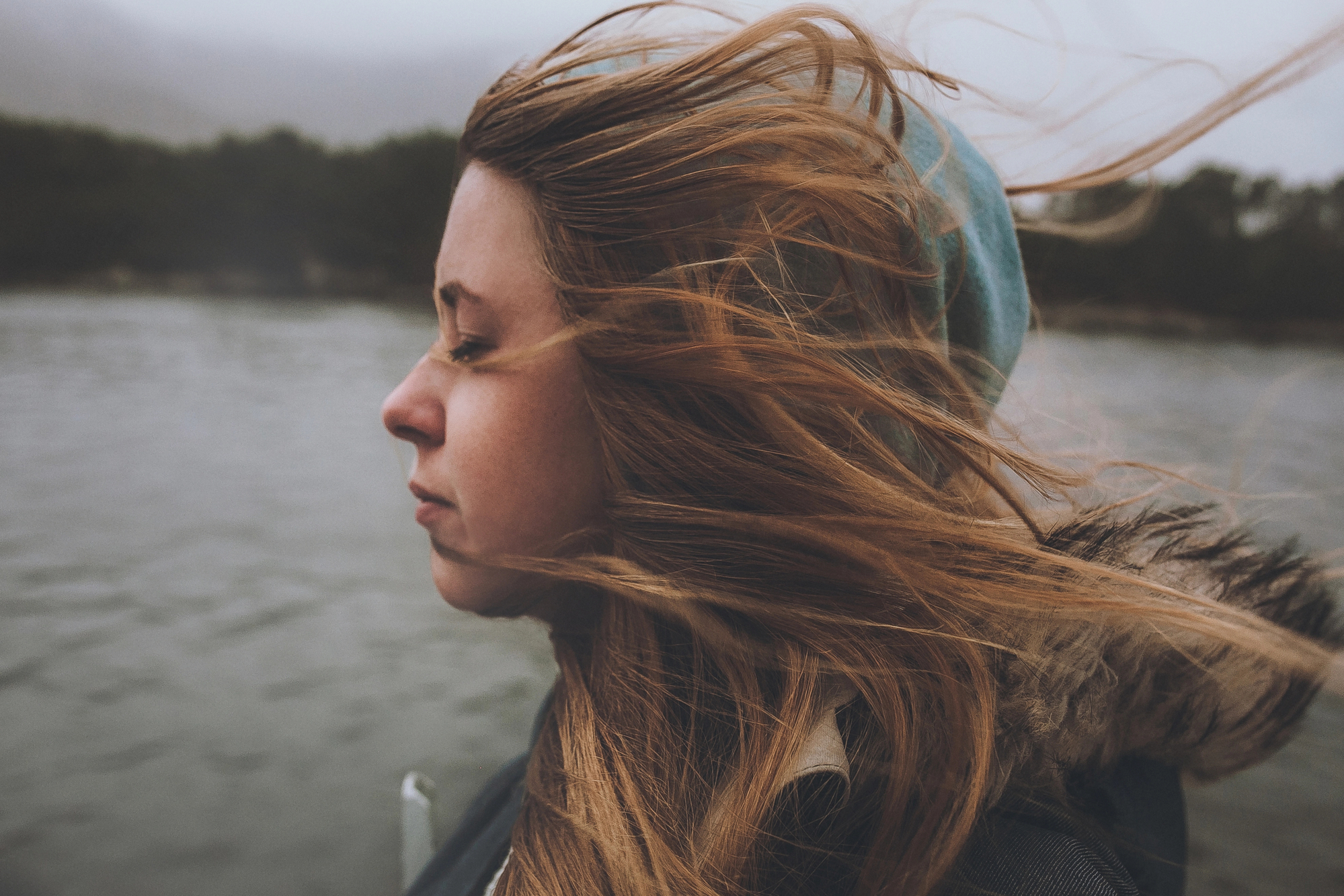 A woman with long hair blowing in the wind stands near a body of water. She is wearing a hooded jacket with fur trim and has her eyes closed, appearing contemplative or peaceful. The background is blurred, with dark foliage and cloudy skies.