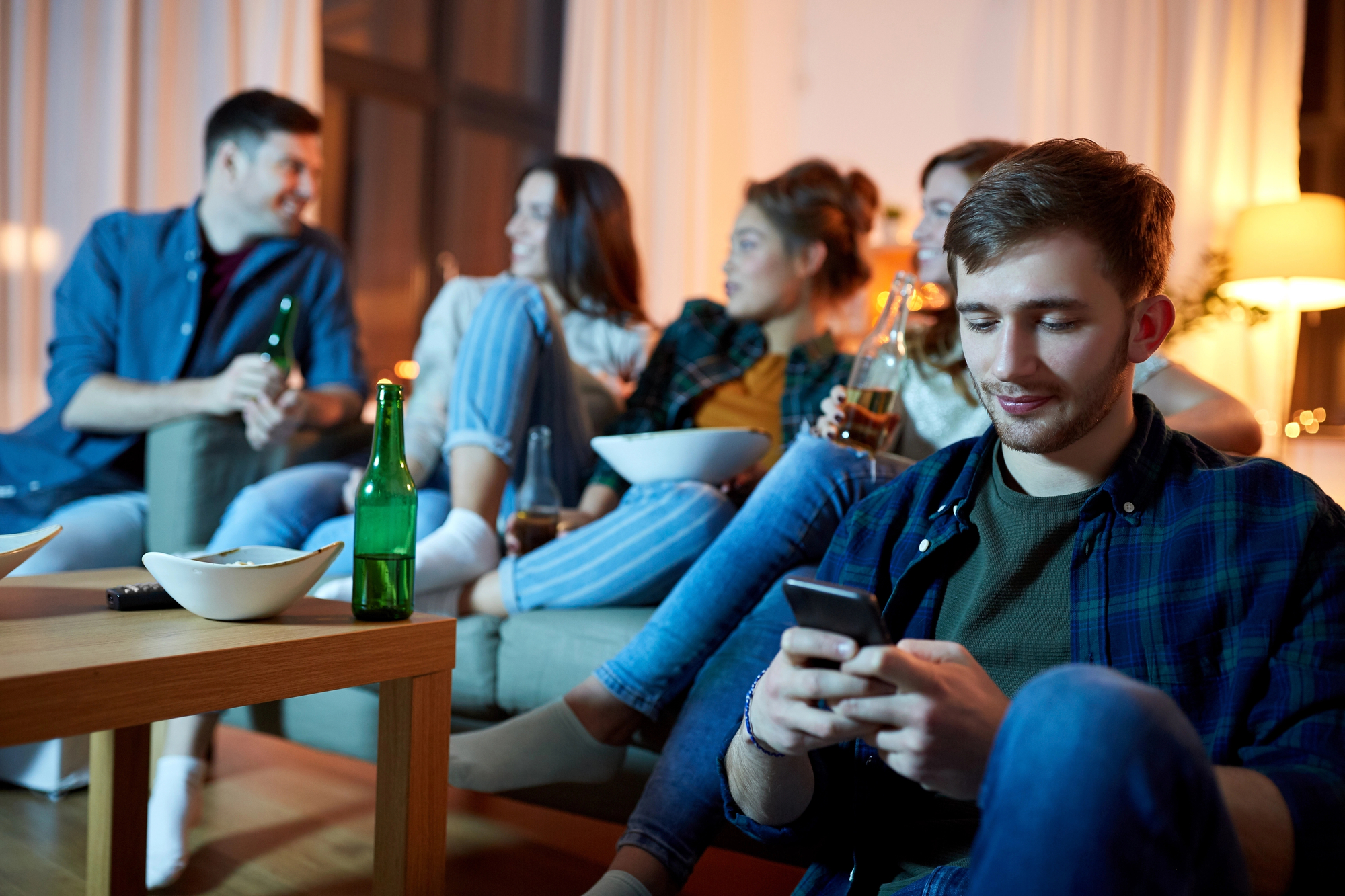 A group of five friends is sitting on a couch in a cozy living room. Four of them are engaged in conversation and holding drinks, while one person in the foreground is focused on their smartphone. The setting includes a coffee table with a couple of bottles and bowls.