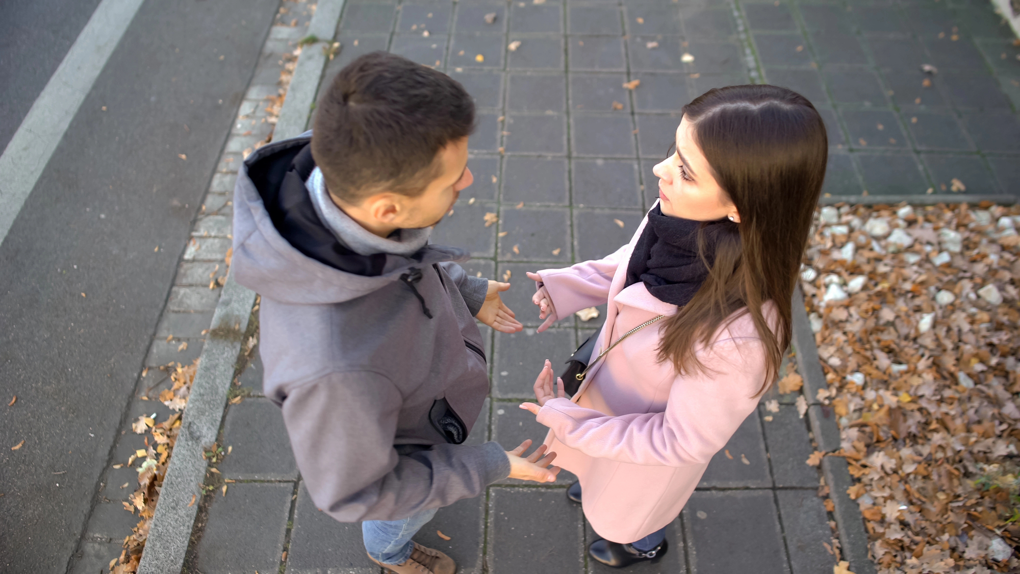 A man and woman are standing on a sidewalk, facing each other and engaged in conversation. The man wears a gray jacket, while the woman is dressed in a pink coat and black scarf. Fallen leaves are scattered on the ground around them.