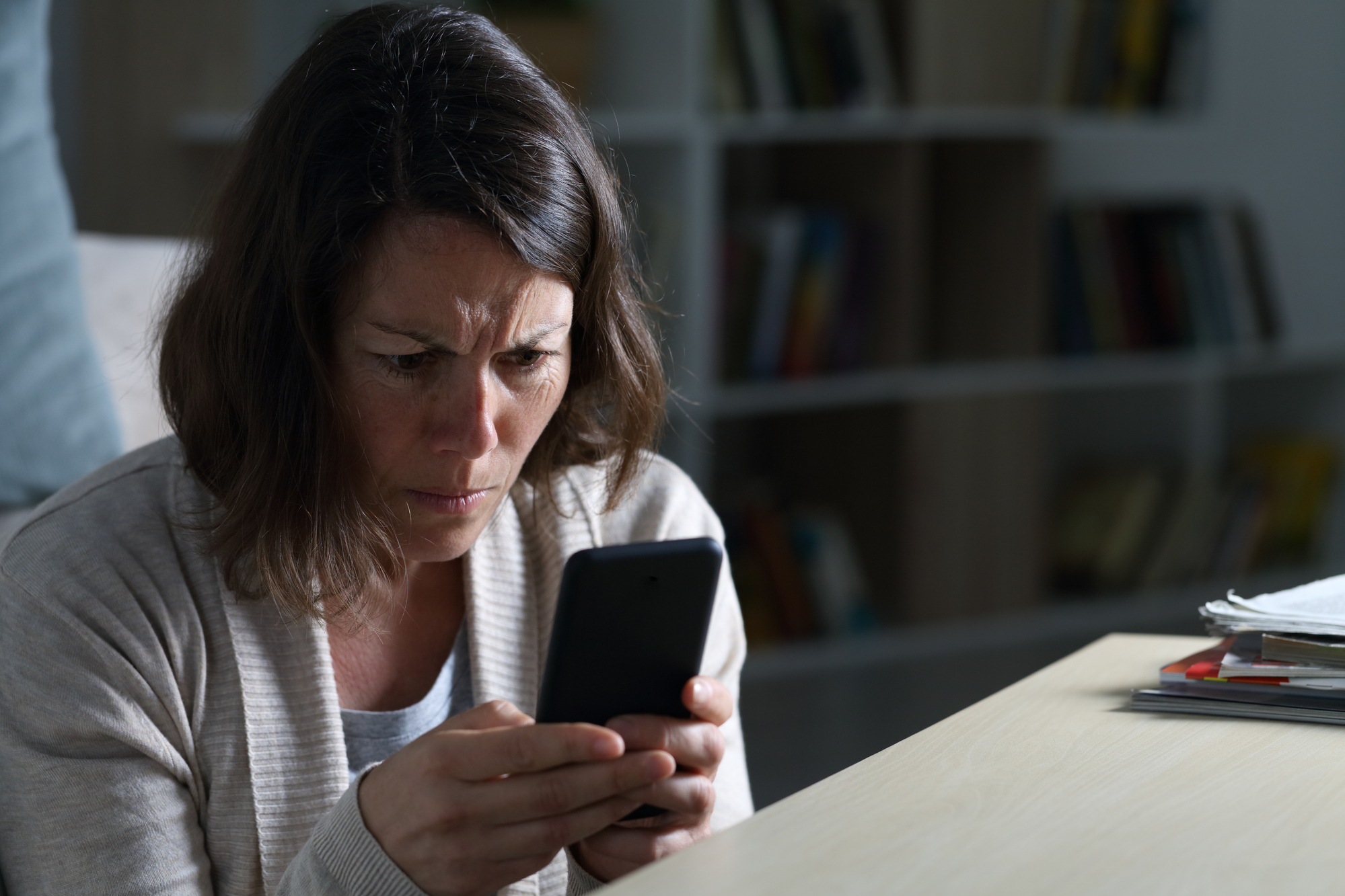 A woman with shoulder-length brown hair concentrates intensely while looking at a smartphone in her hands. She is sitting indoors, with bookshelves in the background. The lighting is dim, creating a serious atmosphere.