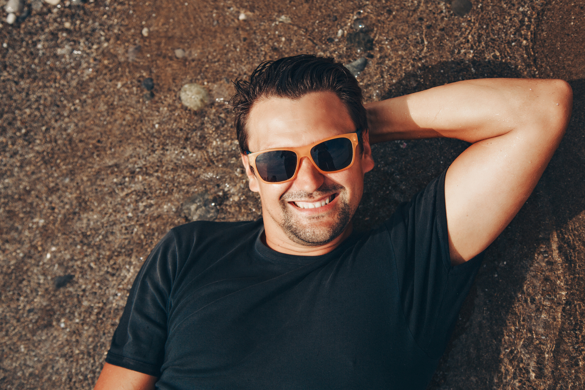 A man with short dark hair is lying on his back on a sandy beach, smiling and wearing orange-framed sunglasses and a black T-shirt. His right arm is bent and resting behind his head. The beach is covered with small pebbles and wet sand.