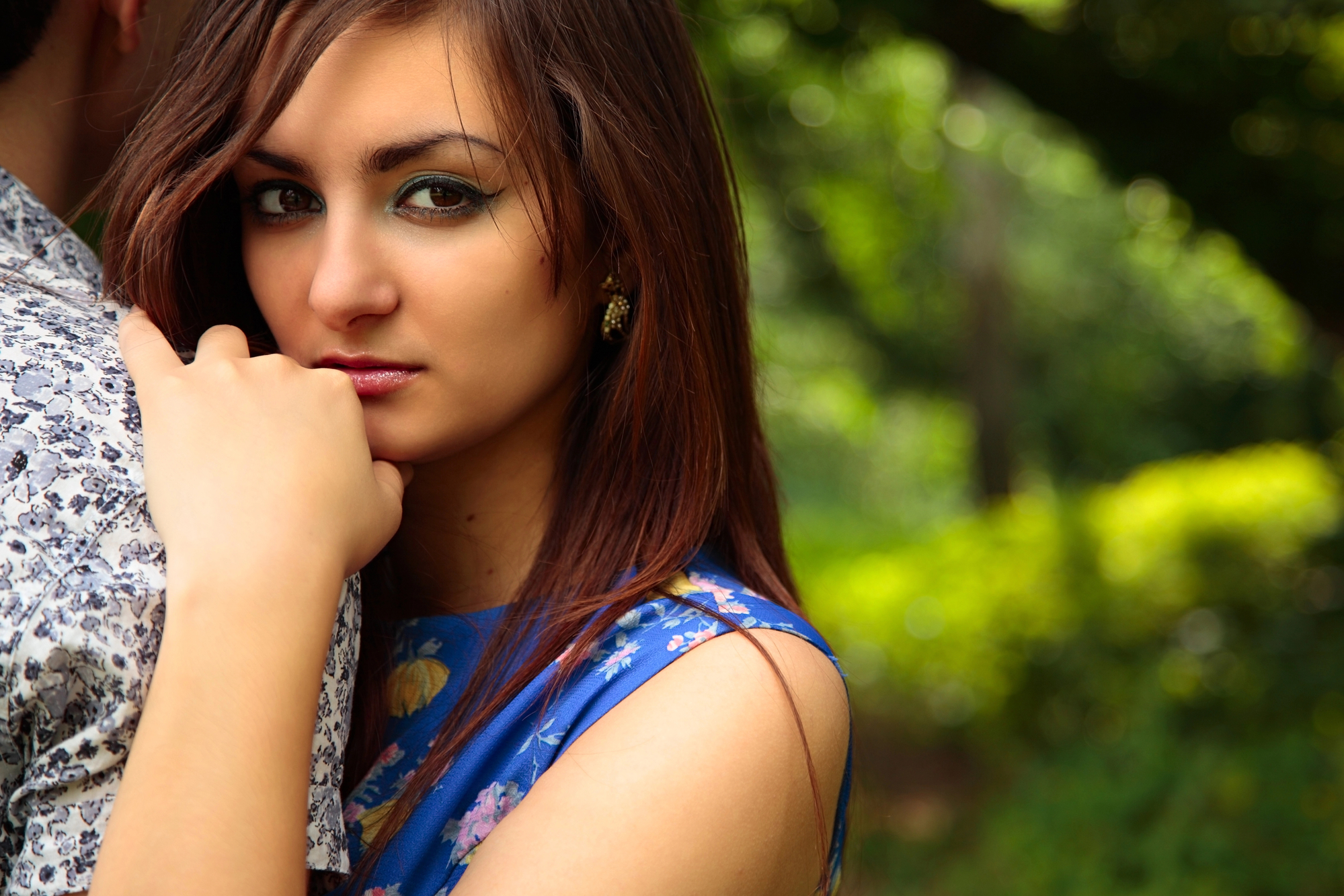 A woman with long brown hair wearing a blue floral dress is standing outdoors. She is resting her hand on her chin and looking directly at the camera. The background is filled with greenery, suggesting a garden or park setting.