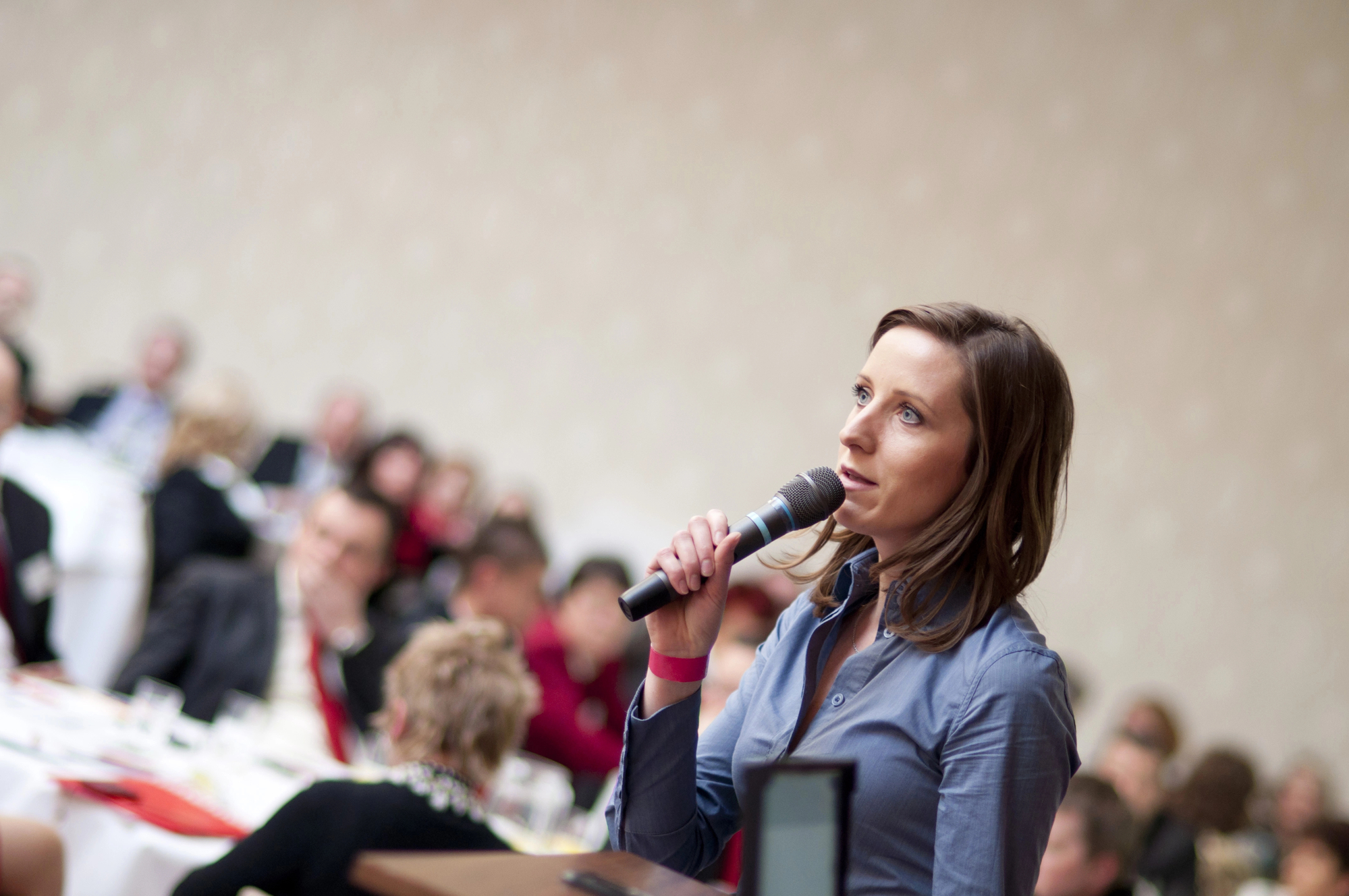 A woman in a blue shirt is speaking into a microphone at a conference. She has light brown hair and is looking thoughtfully ahead. In the background, an audience is seated at round tables, listening attentively. The setting appears to be a formal event or seminar.