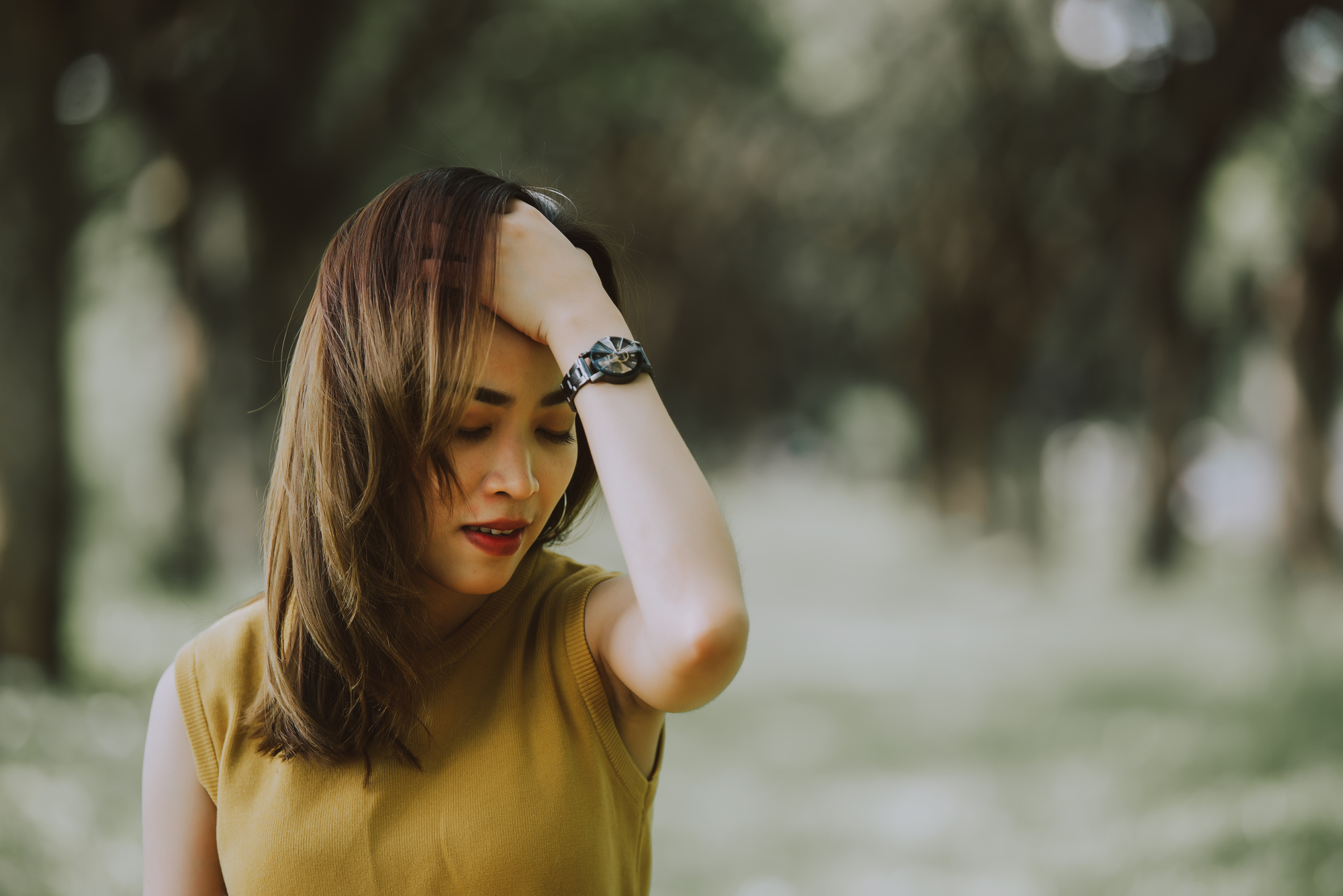 A woman with shoulder-length hair and wearing a sleeveless top is standing outdoors with a blurred background of trees. She is touching her forehead with her hand and appears to be pensive or stressed.