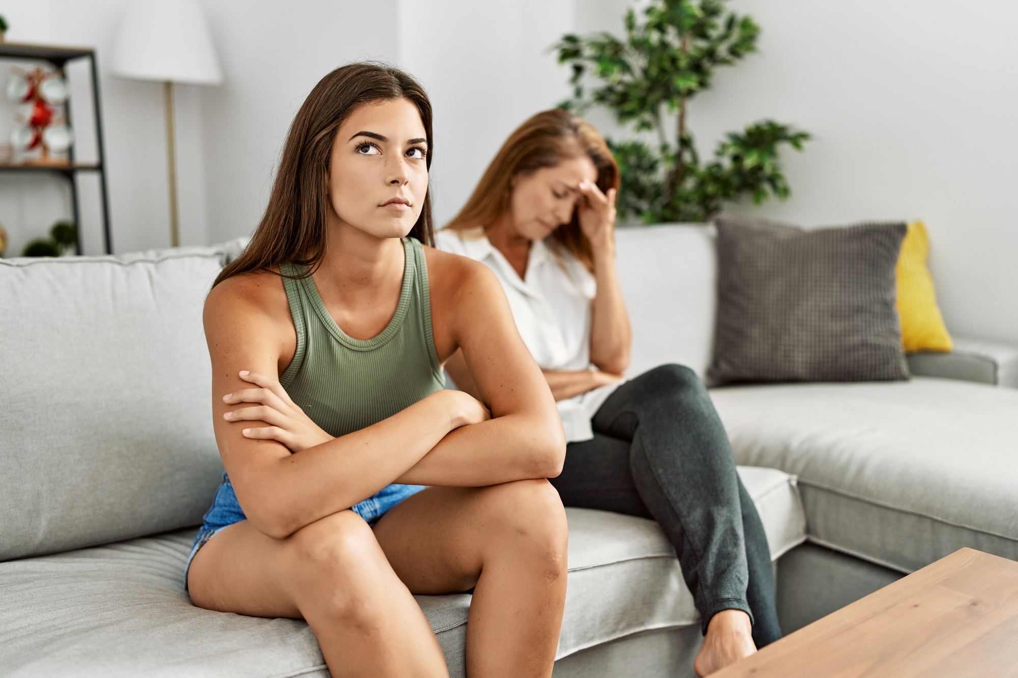 A young woman in a green tank top and denim shorts sits on a couch with her arms crossed, looking away and appearing frustrated. Next to her, an older woman in a white shirt and dark pants sits with her hand on her forehead, looking stressed. A plant is in the background.