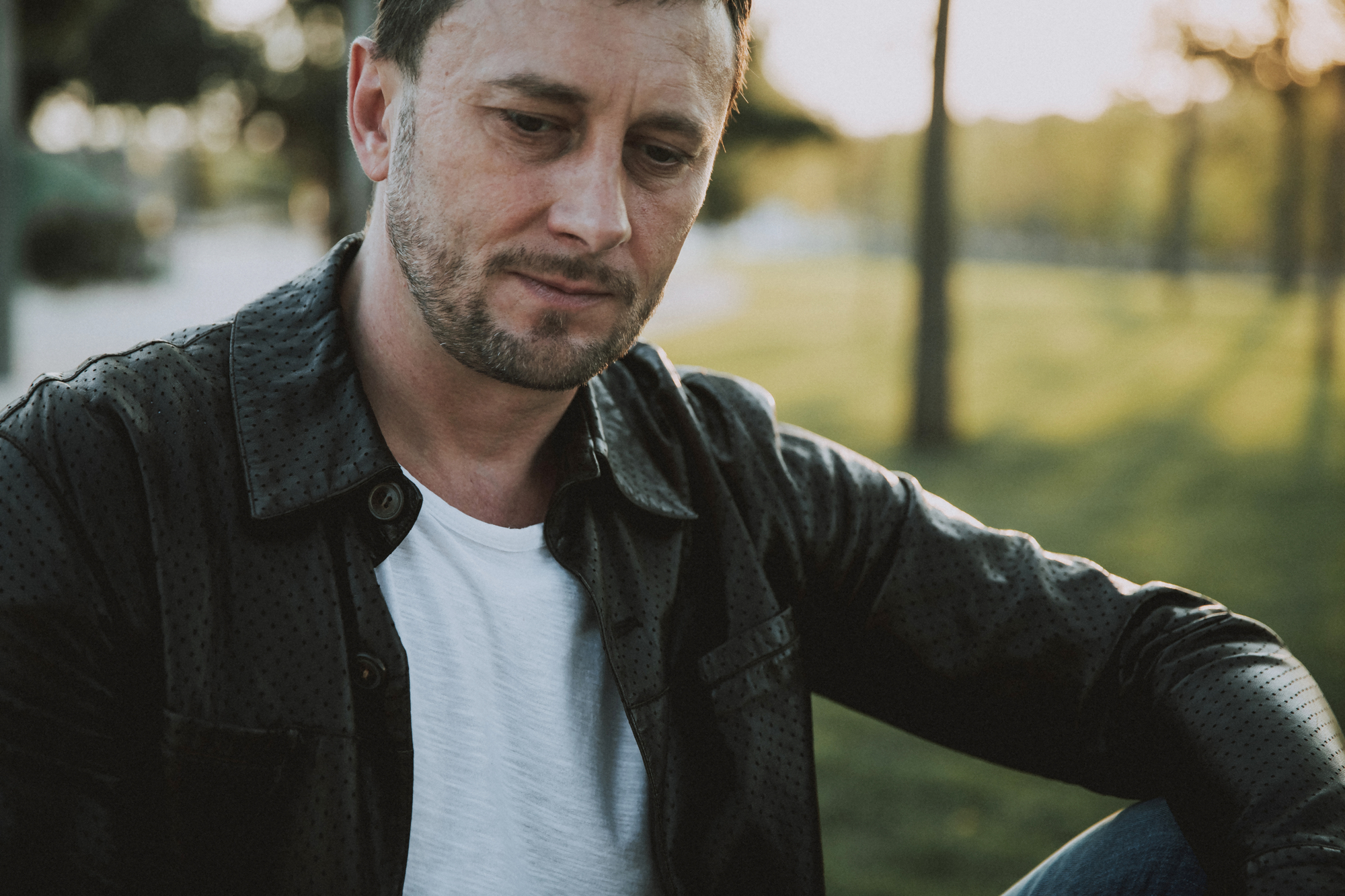 A man in a white t-shirt and dark jacket sits outdoors in a park-like setting, with trees and grass in the background. He is looking down with a thoughtful or contemplative expression. The scene is illuminated by soft, natural sunlight.