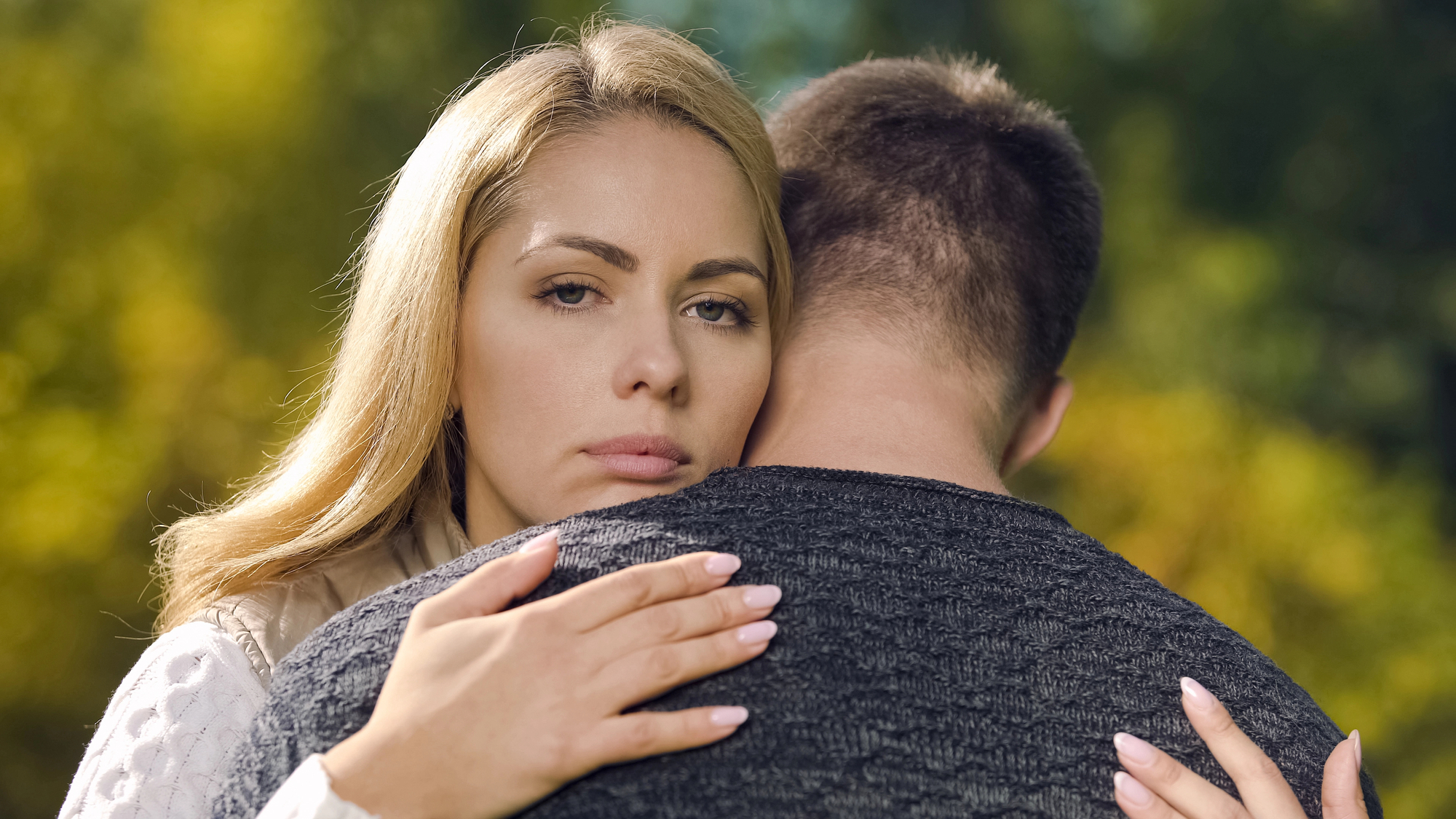 A woman with blonde hair appears serious as she embraces a man whose back is facing the camera. They stand outdoors with a blurred, green natural background.