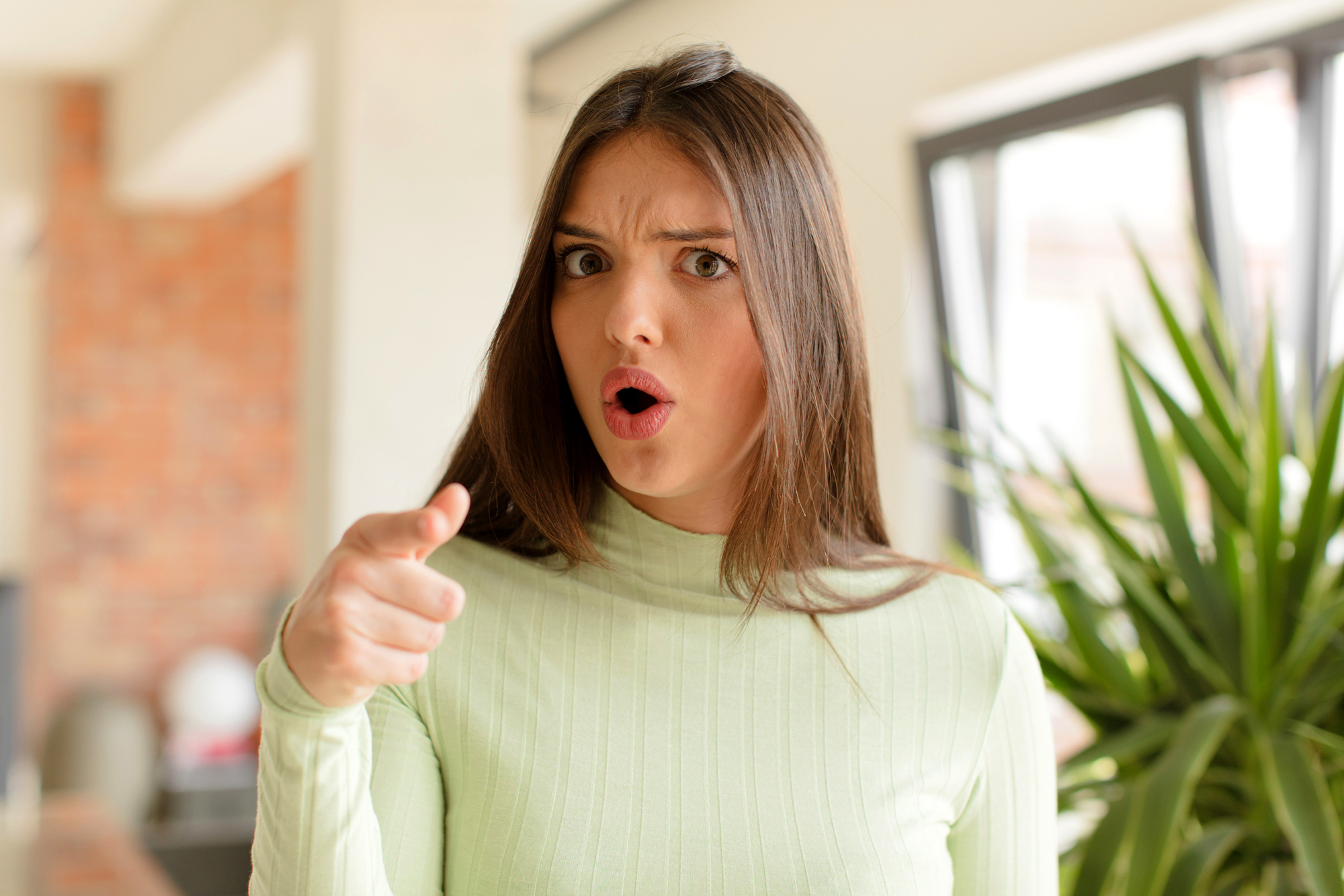 A young woman with long brown hair, wearing a light green turtleneck, looks surprised and points directly at the camera. She stands in a well-lit room with a brick wall and green plant visible in the background.