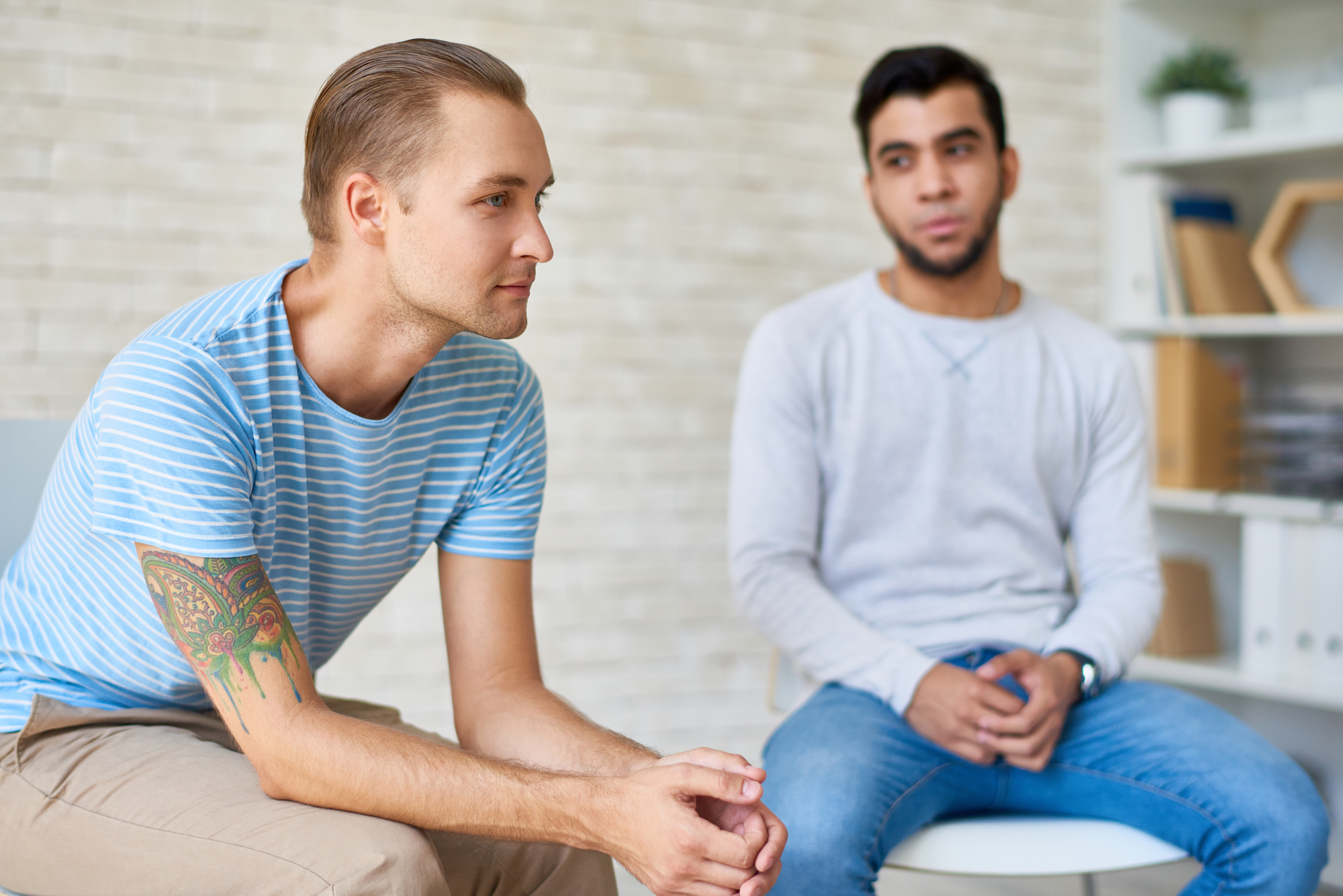 Two men sitting in a light-filled room. The man on the left, wearing a striped t-shirt and sporting a tattoo on his left arm, looks thoughtful. The man on the right, in a long-sleeve shirt and jeans, is looking ahead. Shelves with books are visible in the background.