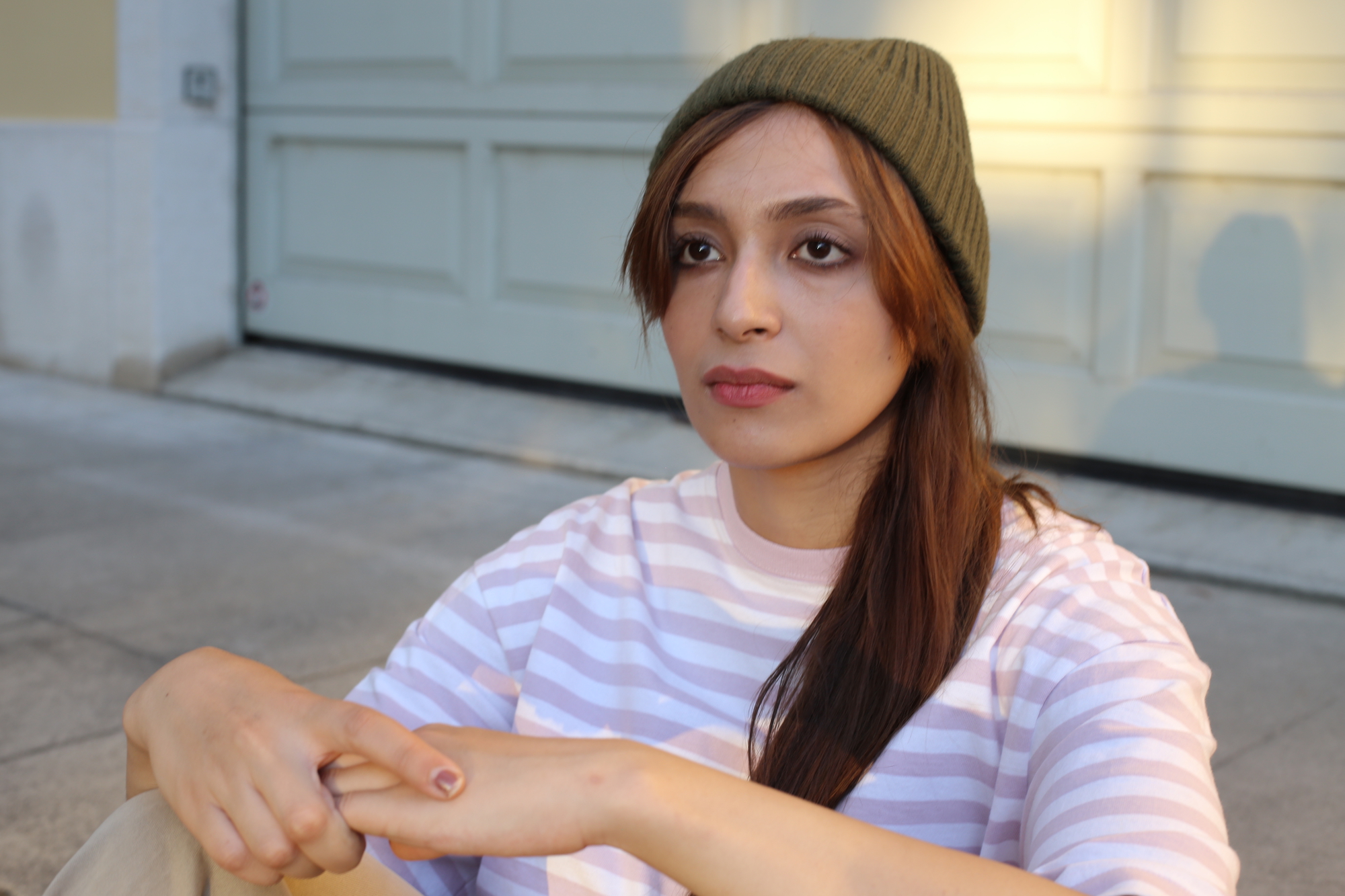 A person with long hair sits on the ground in an outdoor setting. They are wearing a green beanie and a light-colored striped shirt. Their hands are resting on their knees, and they have a thoughtful expression on their face. A light-colored garage door is in the background.
