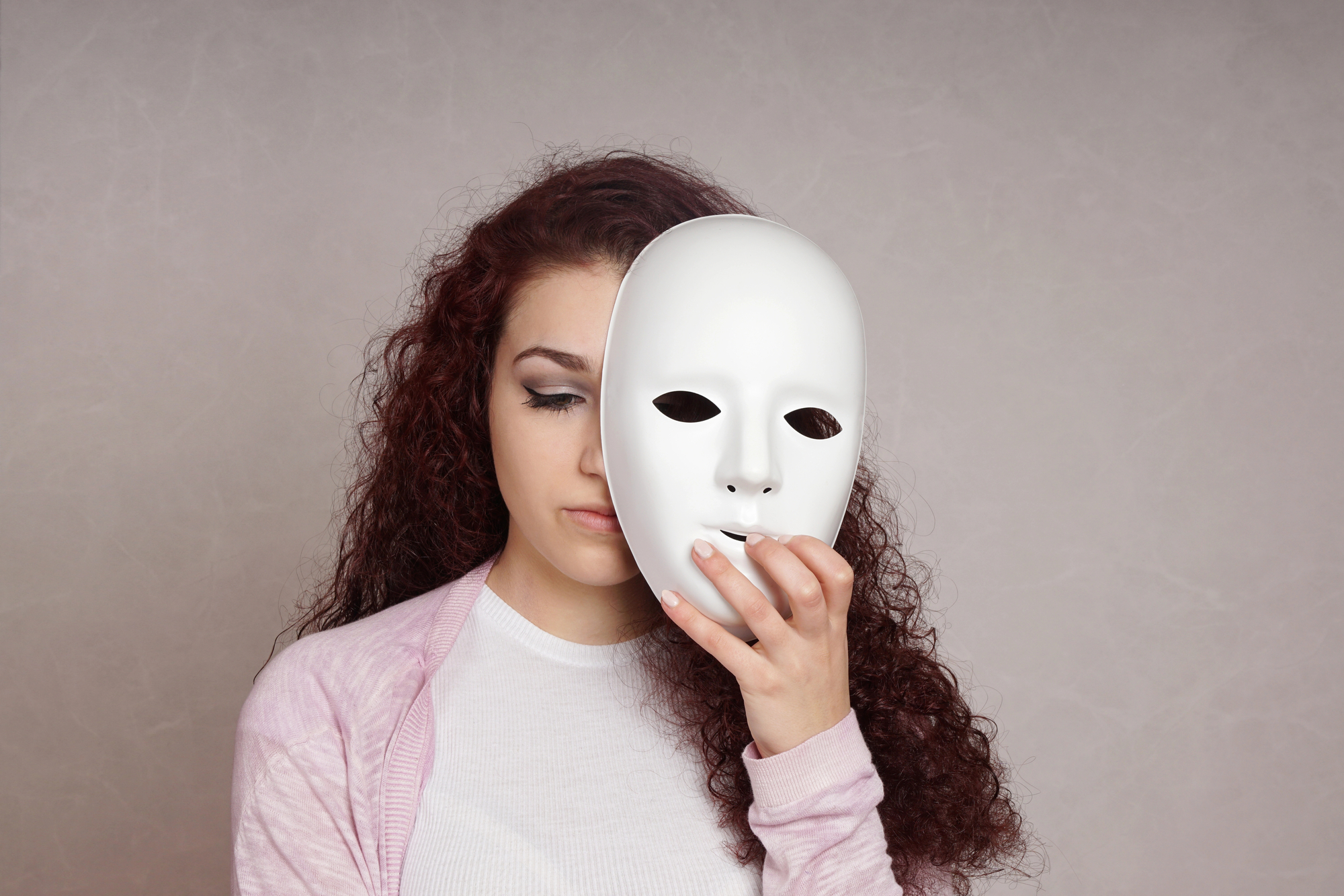 A young woman with long, curly hair holds a white, featureless mask in front of her face, slightly off to one side, revealing her closed eyes. She is wearing a light pink cardigan over a white shirt and stands against a neutral gray background.