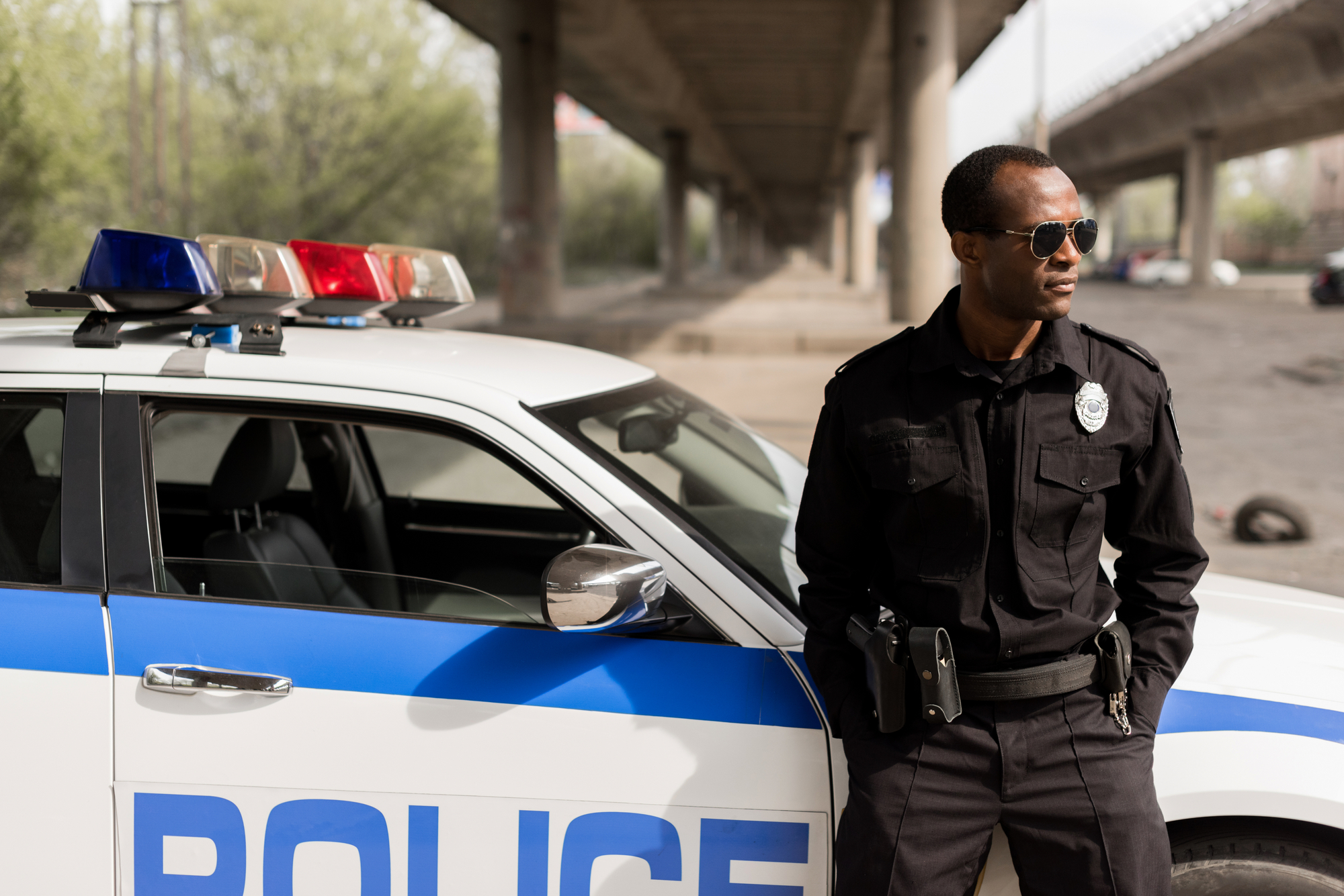 A police officer wearing sunglasses stands next to a police car with blue stripes and a light bar on top. The officer is under a bridge and has one hand resting on the car. Trees and an urban environment are visible in the background.