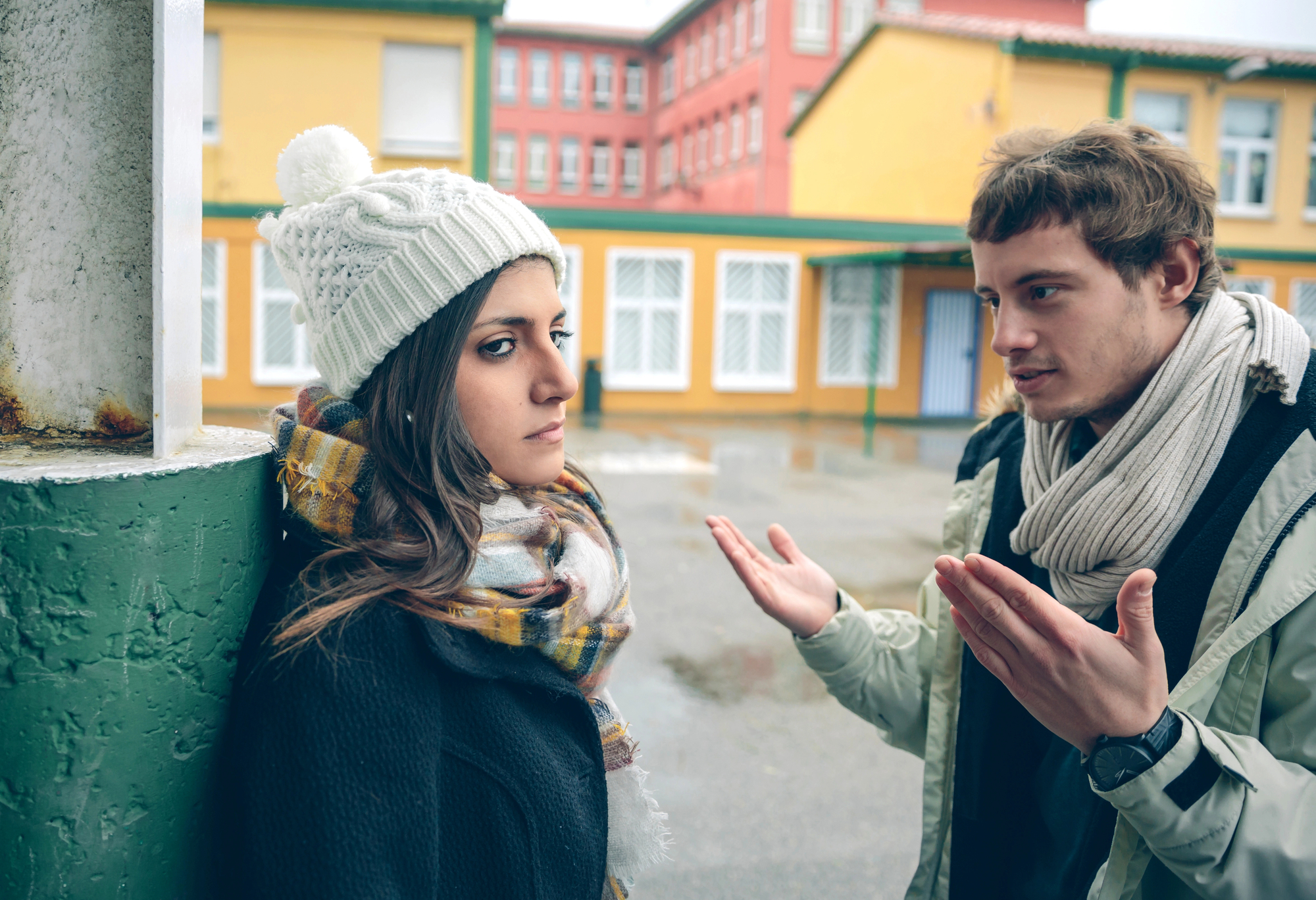 Two people are outside, engaged in a serious conversation. One person, wearing a white beanie and a scarf, leans against a green pillar and looks away. The other person, wearing a scarf and gesturing with both hands, speaks animatedly. They are in a schoolyard area.