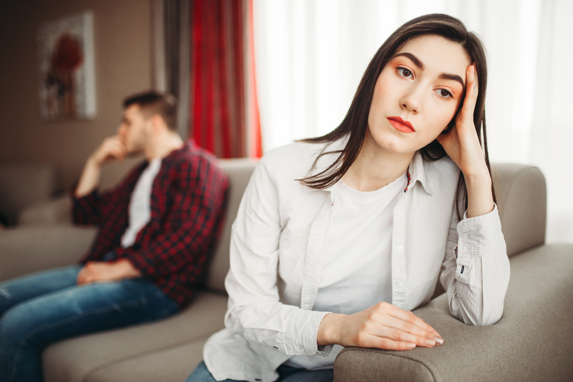 A woman with a thoughtful and concerned expression sits on a couch, resting her head on her hand. In the background, a man sits on a separate couch looking away, seeming distant. The setting appears to be a living room with muted tones and a bright window.