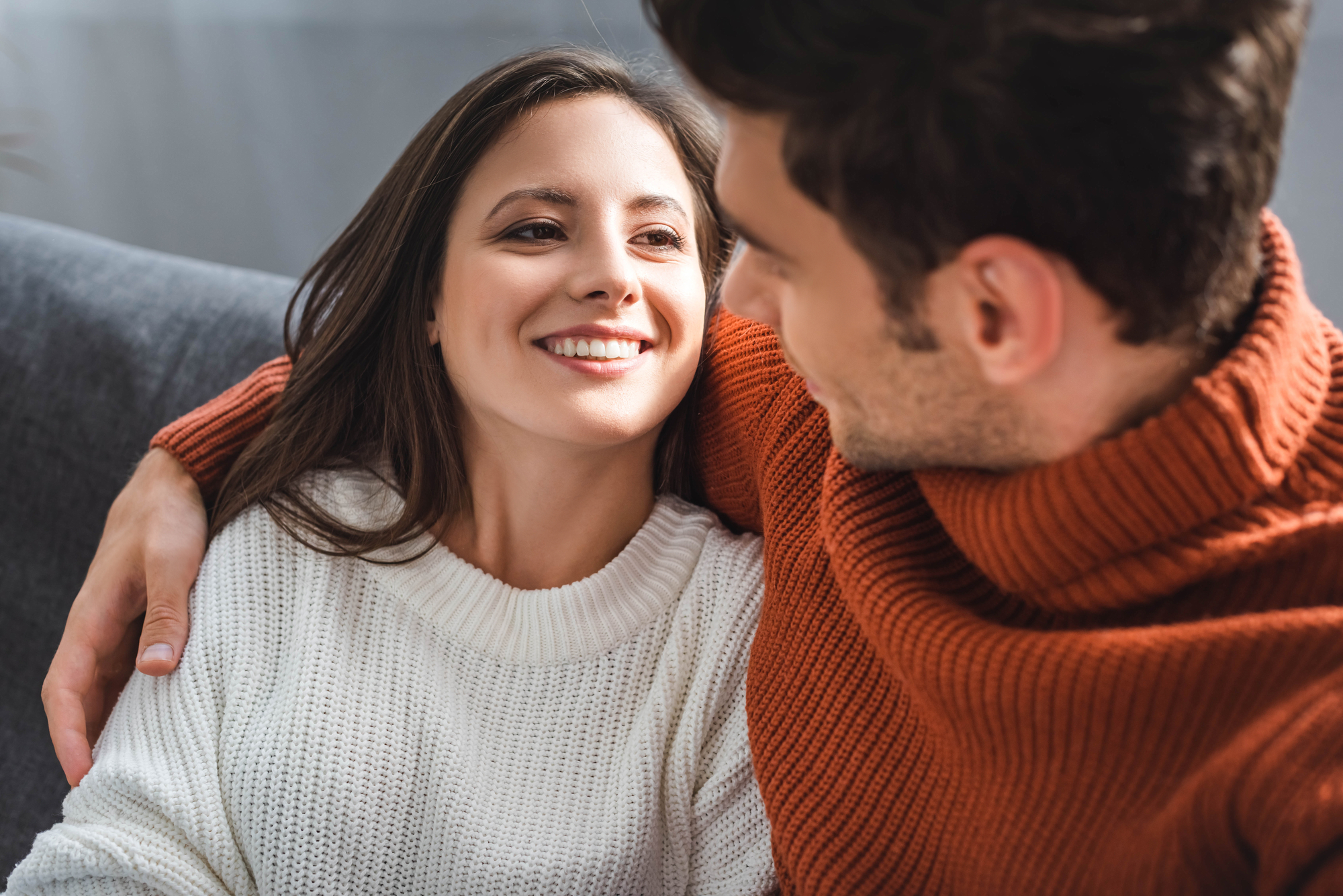 A smiling couple sits closely together on a couch. The woman, wearing a white sweater, looks happily at the man, who is in a rust-colored sweater with his arm around her. They appear to be enjoying a tender moment.