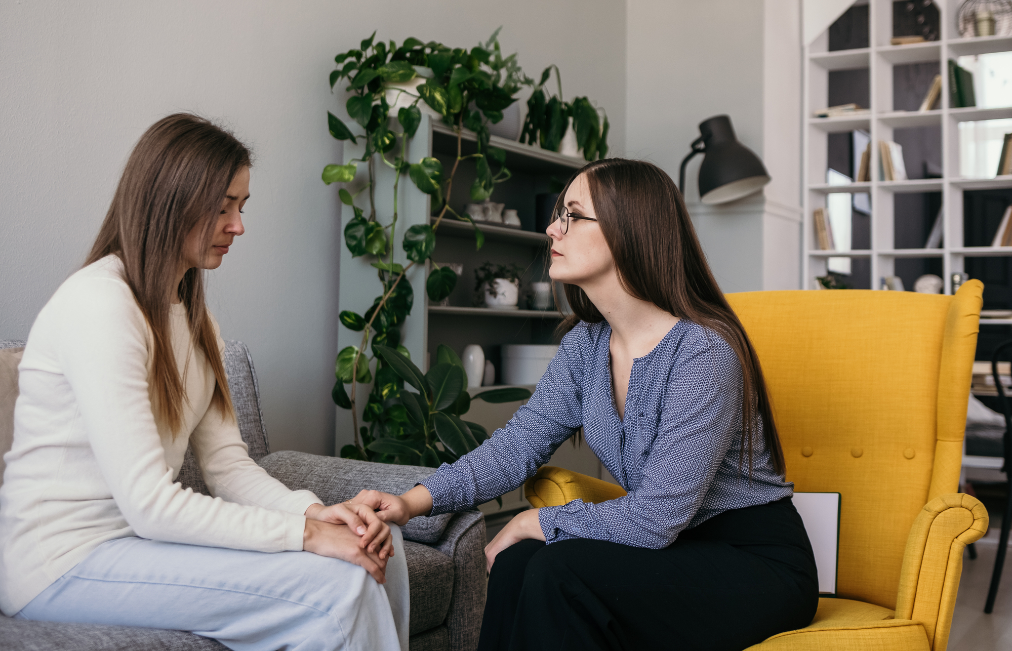 Two women are sitting in a room; one appears supportive, gently holding the other's hand. One woman, in a blue blouse and glasses, sits on a yellow chair, while the other, in a white sweater, sits on a gray chair. Potted plants and shelves are in the background.