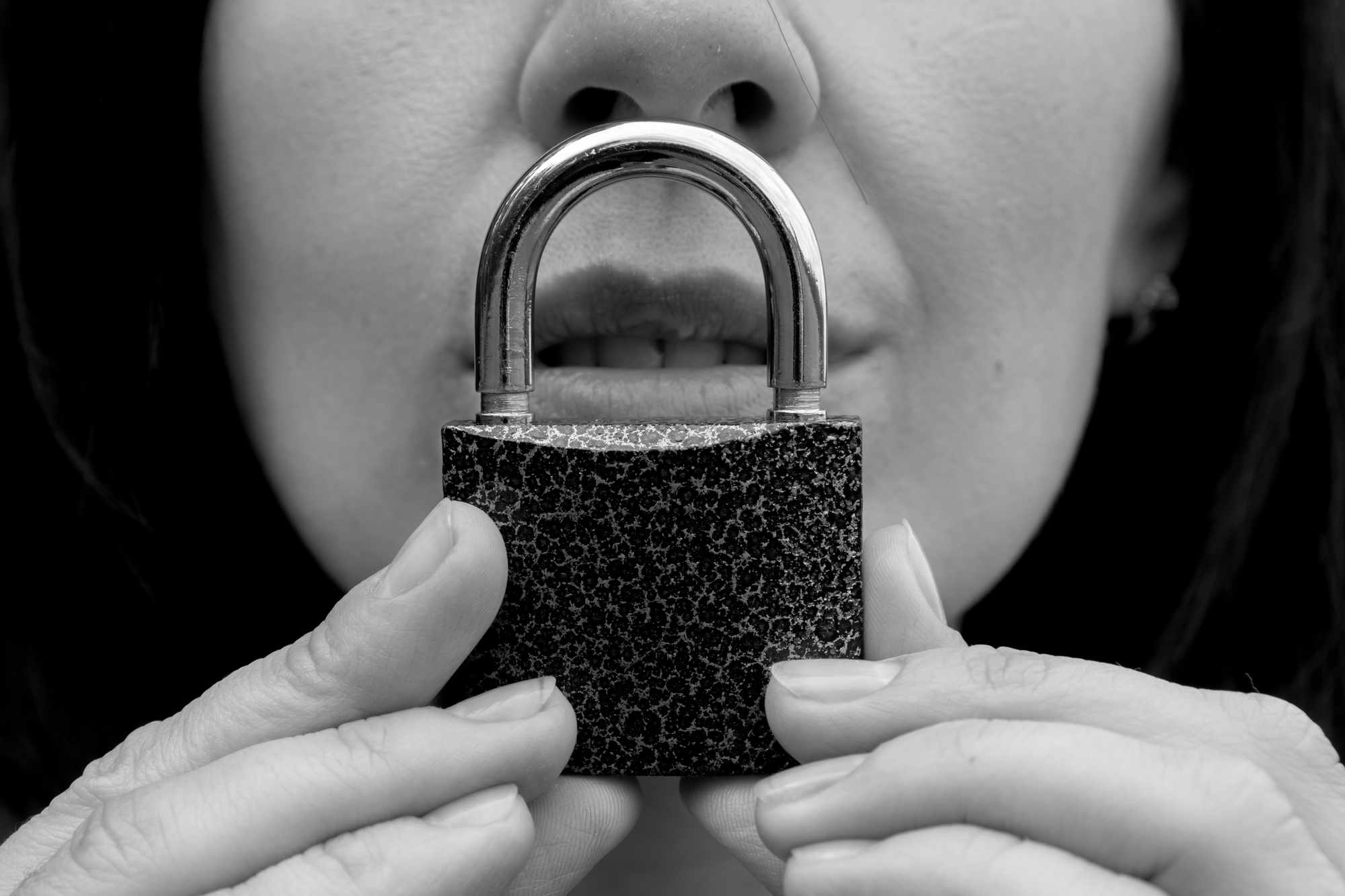 A person holds a padlock closely to their partially visible mouth, symbolizing silence or secrecy. The image is in black and white, focusing on the padlock and the lower part of the person's face.