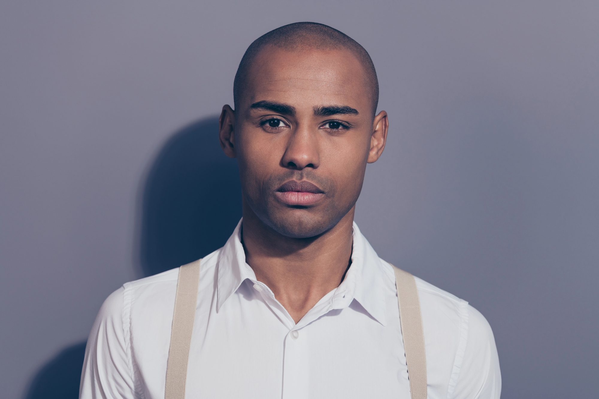 A man with a serious expression stands against a gray background. He is wearing a white dress shirt with beige suspenders and has a bald head. The lighting casts a soft shadow on the wall behind him.