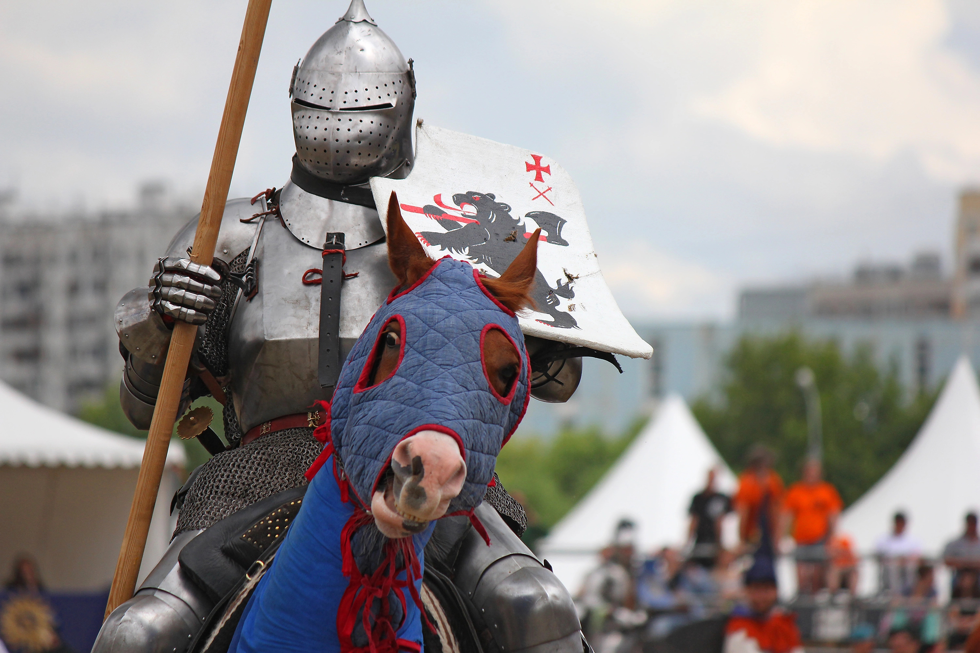 A medieval knight in full armor holds a lance and a shield with a dragon emblem while riding a horse. The horse is draped in a blue and red cover. In the background, there are modern buildings and tents, and spectators are visible.