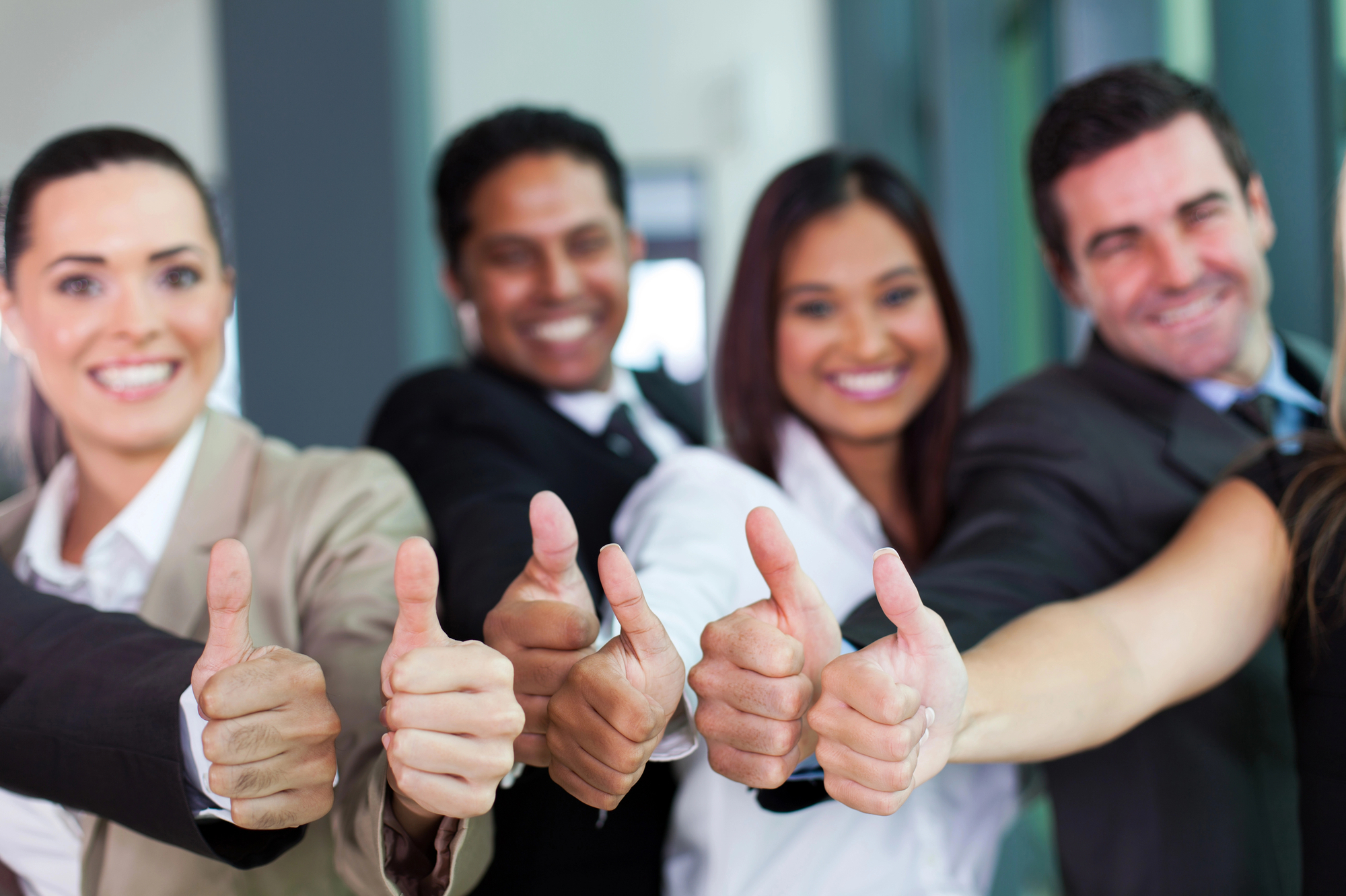 Four people in professional attire, three women and one man, stand in a row with smiles, each giving a thumbs-up. They are in an indoor setting, possibly an office, suggesting a positive, collaborative, or successful work environment.