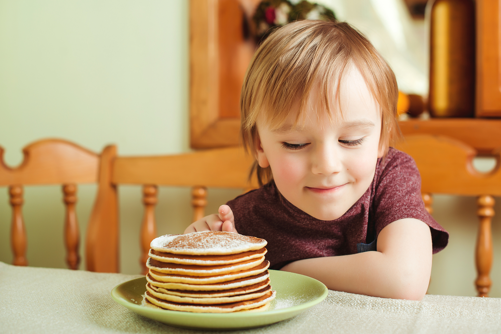 A young child with blonde hair sits at a table, smiling and looking at a stack of pancakes on a green plate in front of them. The child is wearing a maroon shirt, and the background features a wooden chair and a blurred interior setting.