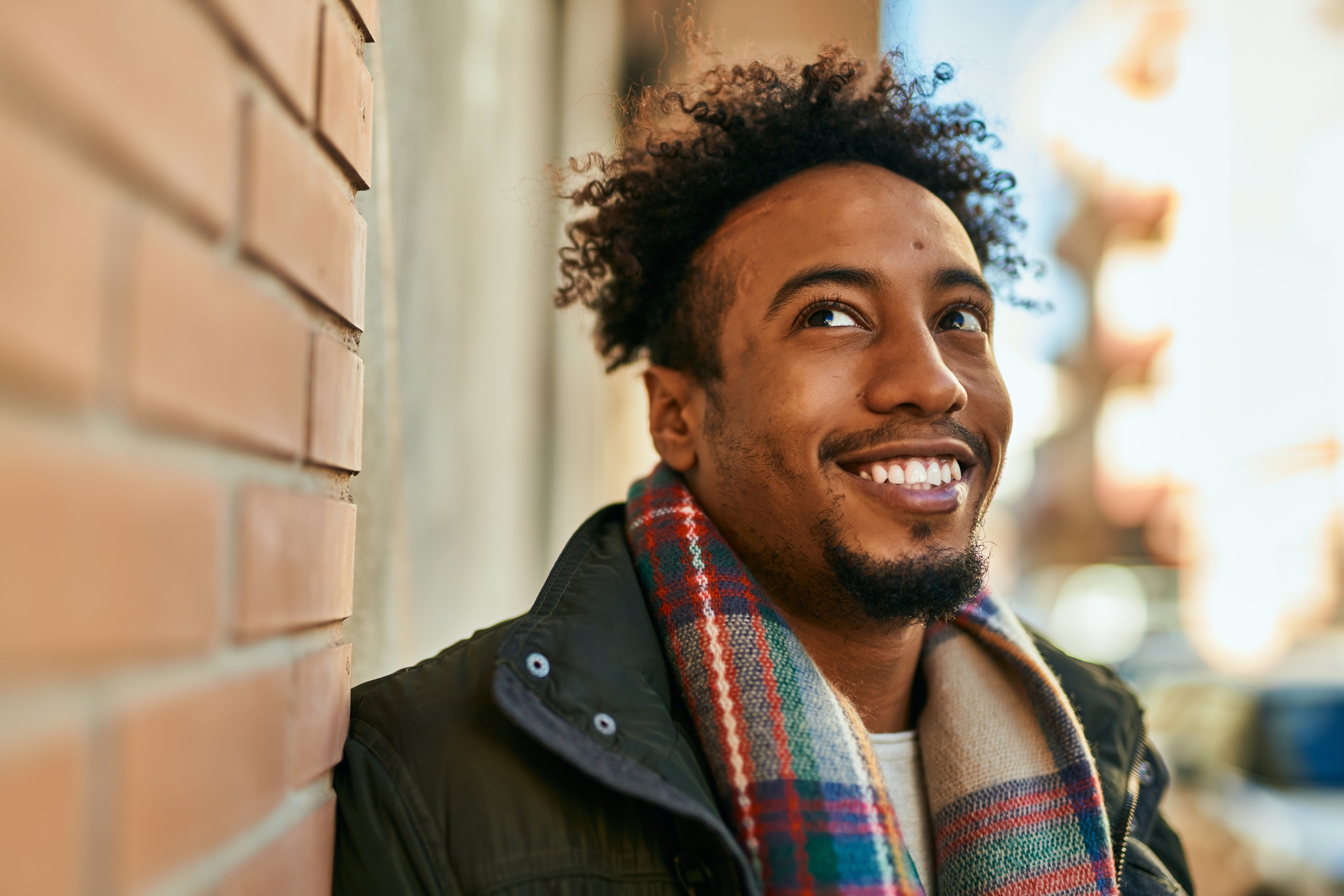 A man with curly hair, a beard, and a mustache smiles warmly while leaning against a brick wall. He is wearing a dark jacket and a colorful plaid scarf. The background shows an outdoor urban setting on a pleasant sunny day.