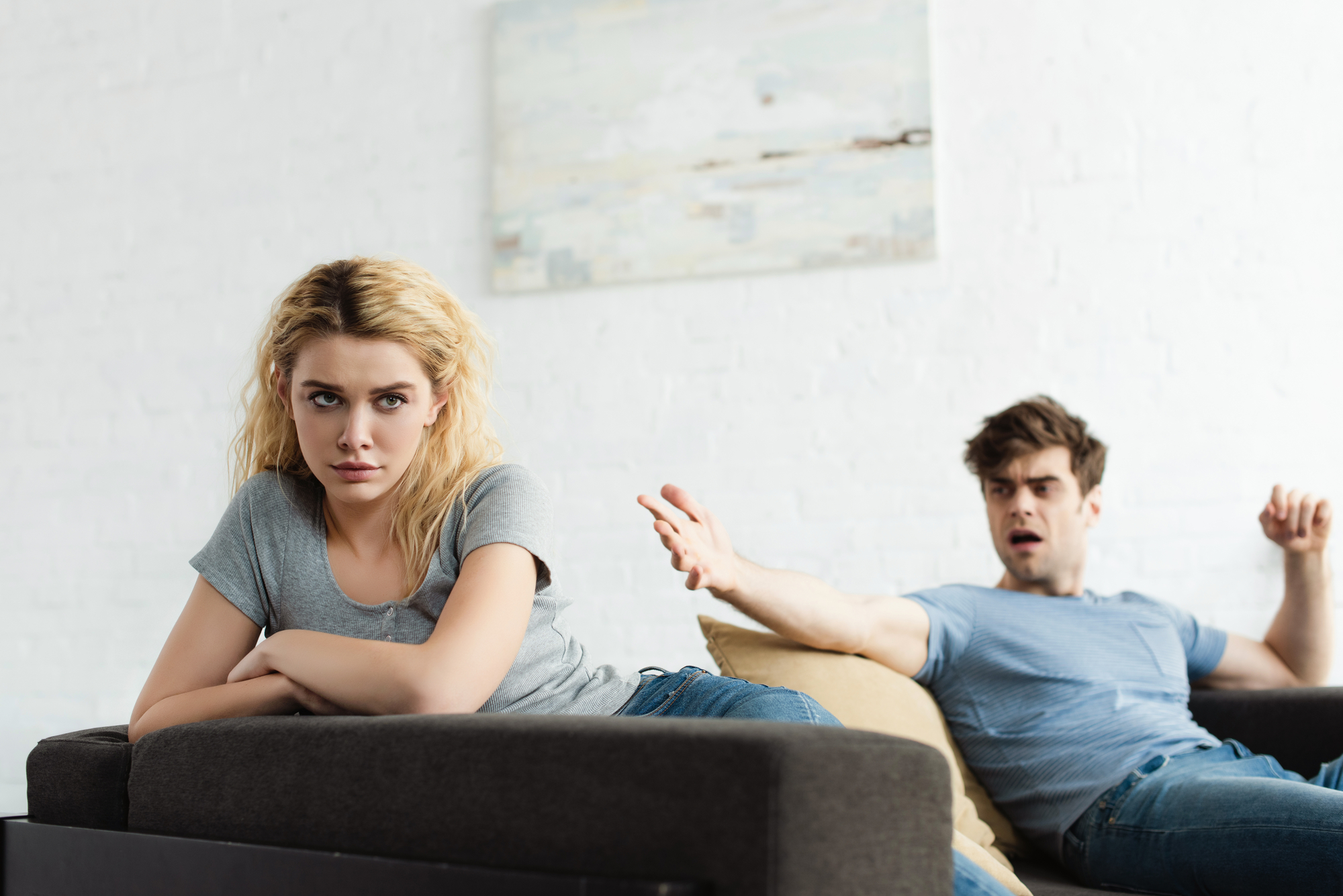A woman with blond hair sits on a couch looking upset, her back turned to a man with dark hair who is seated on an adjacent couch, appearing frustrated with his arm outstretched. The background includes a white wall with a painting.
