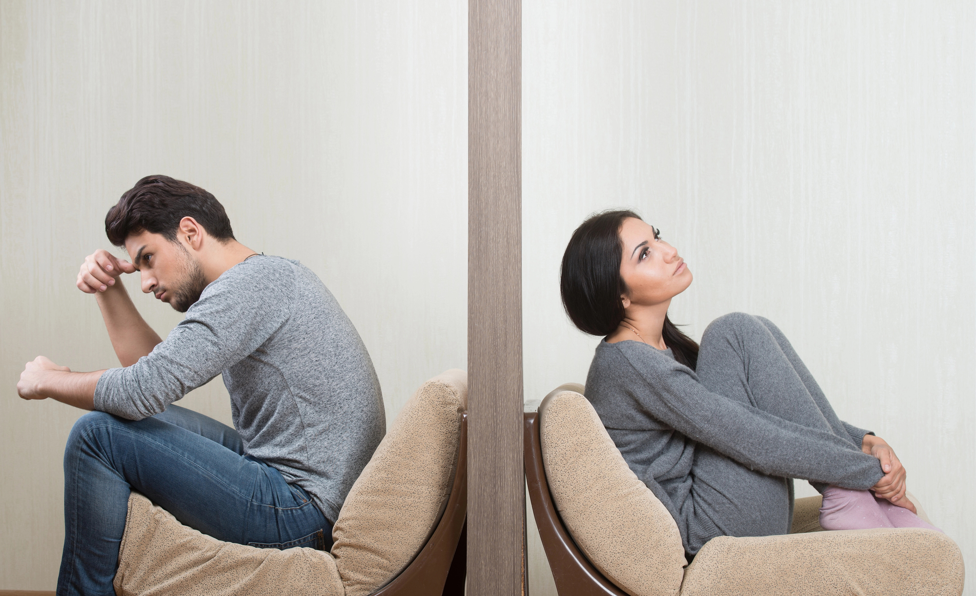 A man and a woman sitting back to back on separate chairs, divided by a wall. The man is leaning forward with his head resting on his hand, appearing stressed. The woman is hugging her knees, looking upwards with a thoughtful expression. Both are dressed in casual clothes.