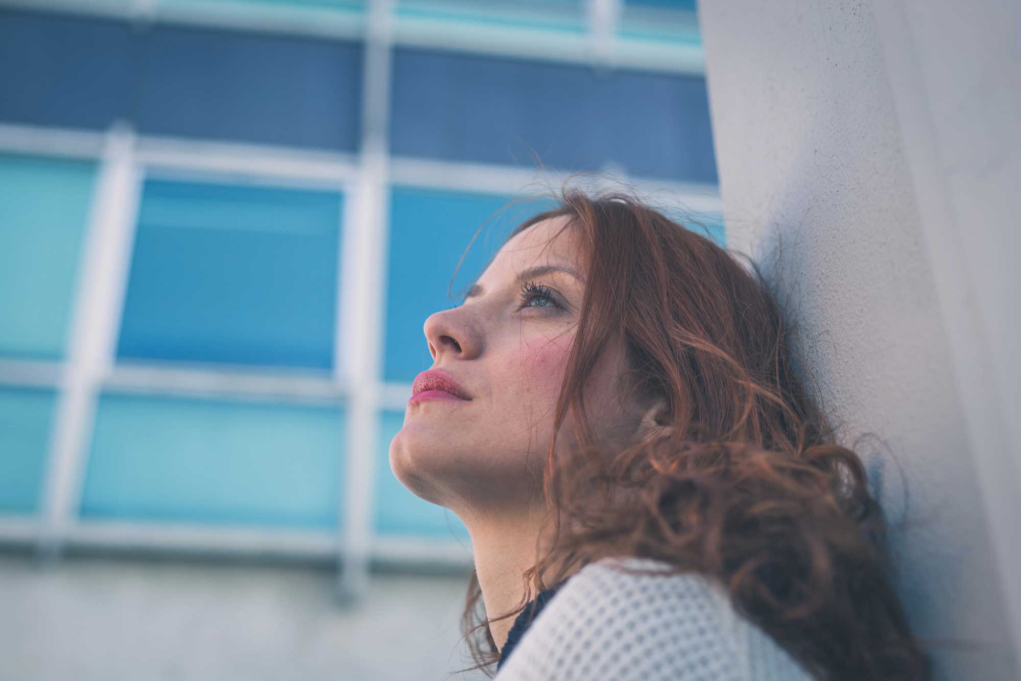 A woman with long, reddish-brown hair leans against a light-colored wall, looking upward with a thoughtful expression. She is outdoors, with the blurred background featuring a modern building with blue-tinted windows.