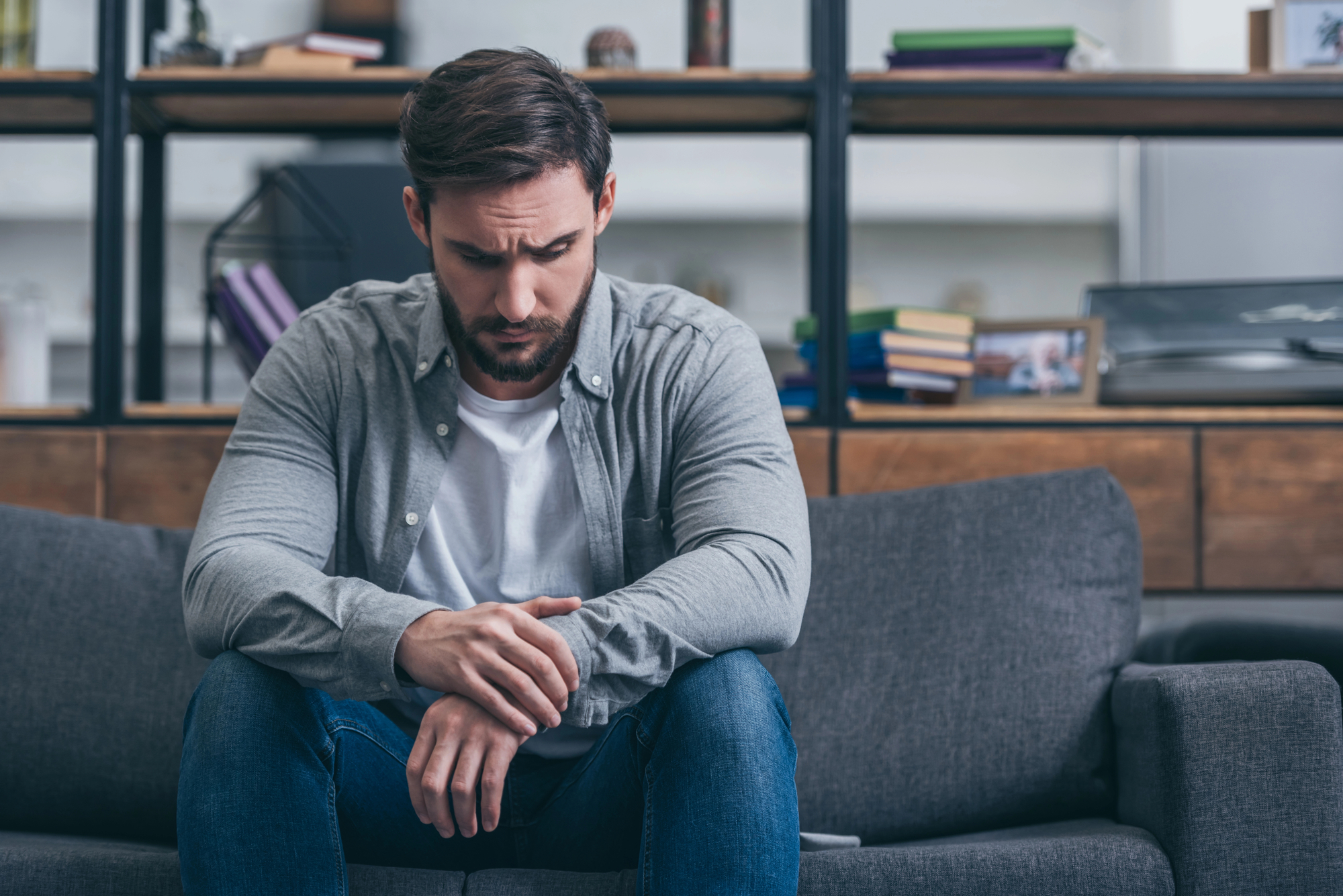 A man with short brown hair and a beard, wearing a gray shirt and blue jeans, sits pensively on a gray sofa with his hands clasped. He is in a modern living room with shelves full of books and decorative items in the background.