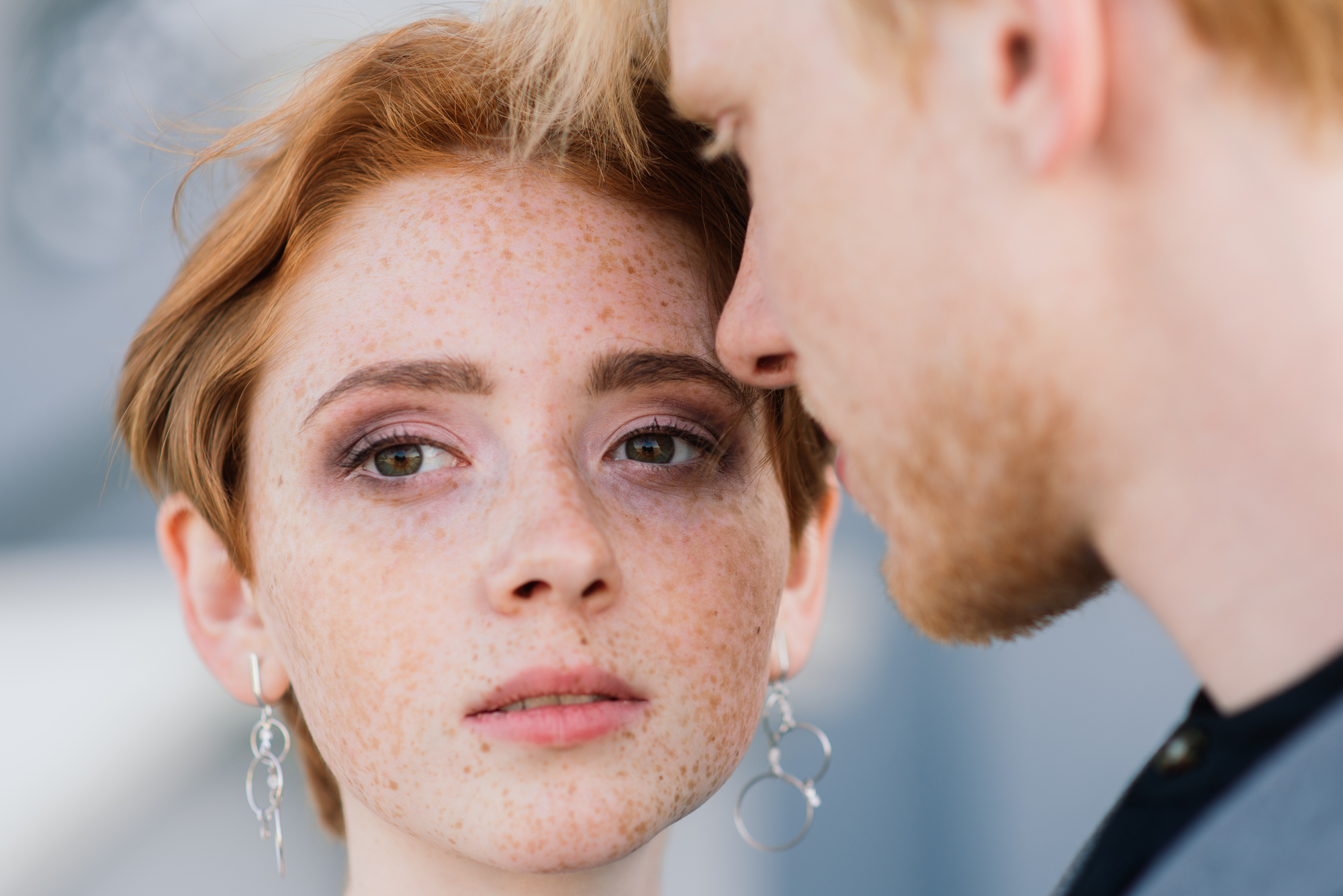 A young woman with short red hair and freckles gazes directly at the camera, wearing silver hoop earrings. Beside her, a man with blonde hair is in profile, appearing to be close to her face. The background is blurred and neutral-toned.