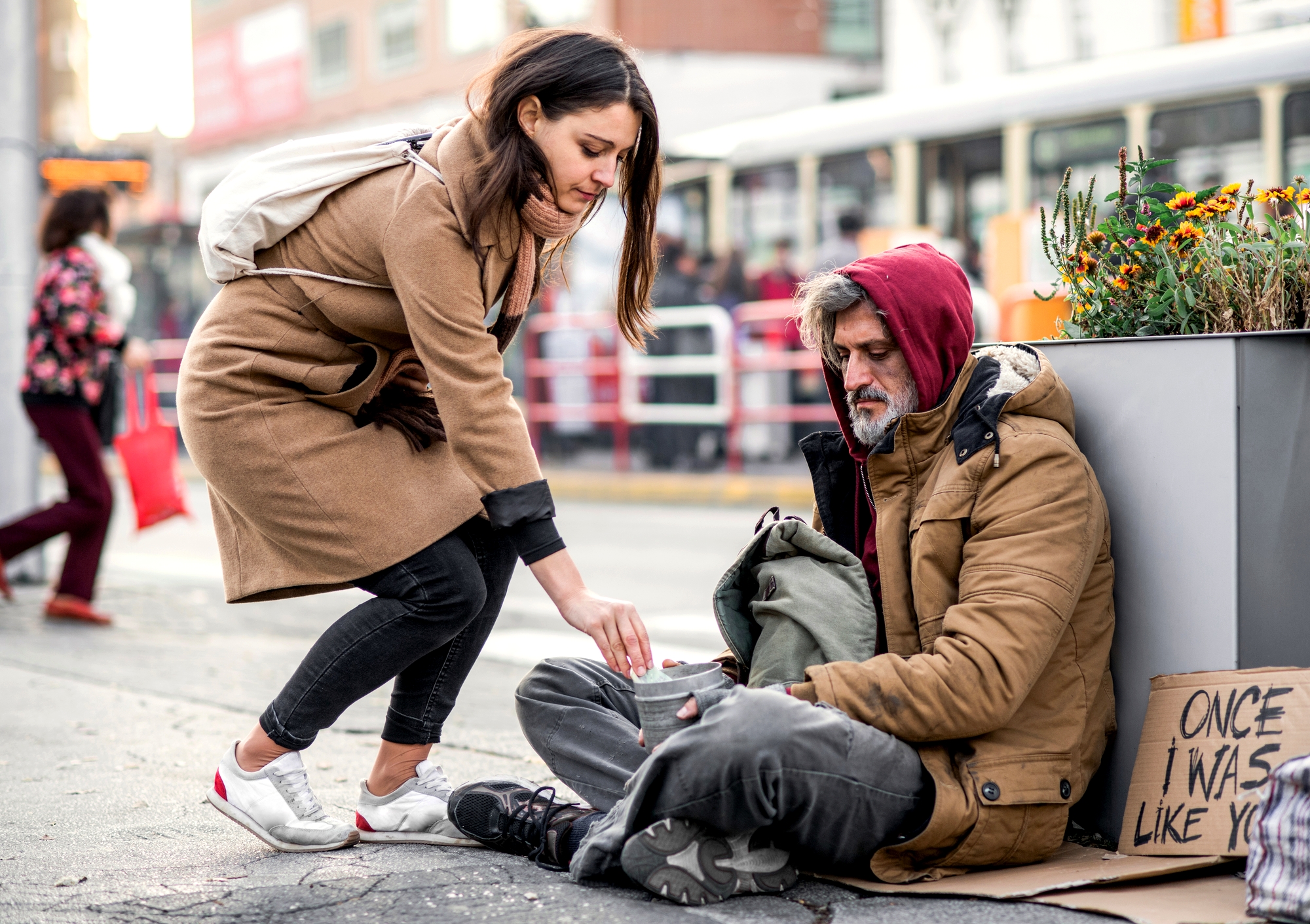 A woman wearing a brown coat and a backpack is bending down to offer change to a homeless man sitting on the sidewalk. The man is bundled in a red hoodie and a brown jacket, holding a cup. A handwritten sign beside him reads, "ONCE I WAS LIKE YOU.
