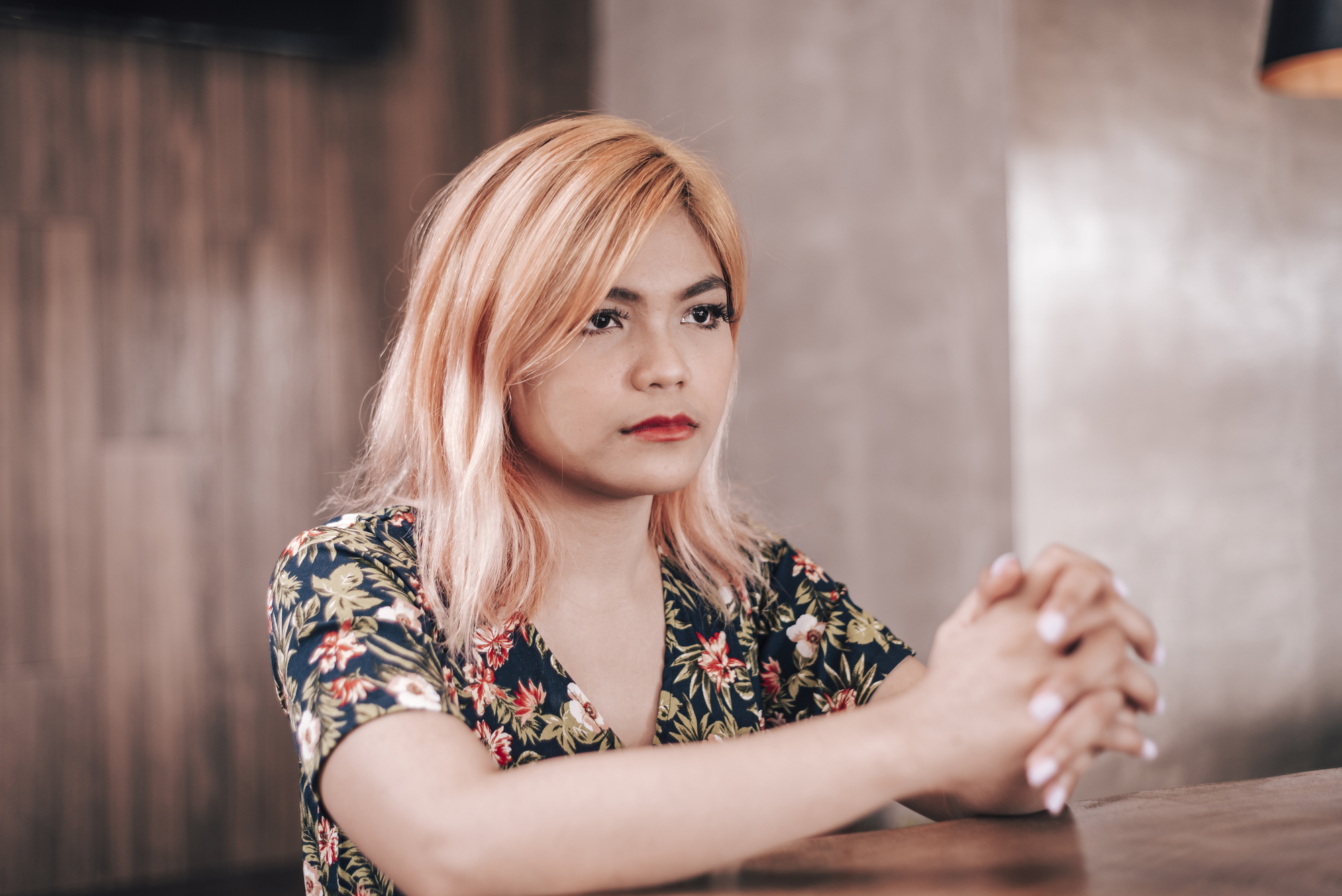 A person with shoulder-length blonde hair and a serious expression is seated at a wooden table. They are wearing a floral patterned shirt and have their hands clasped together, looking ahead thoughtfully. The background features a blurred, warm-toned interior.