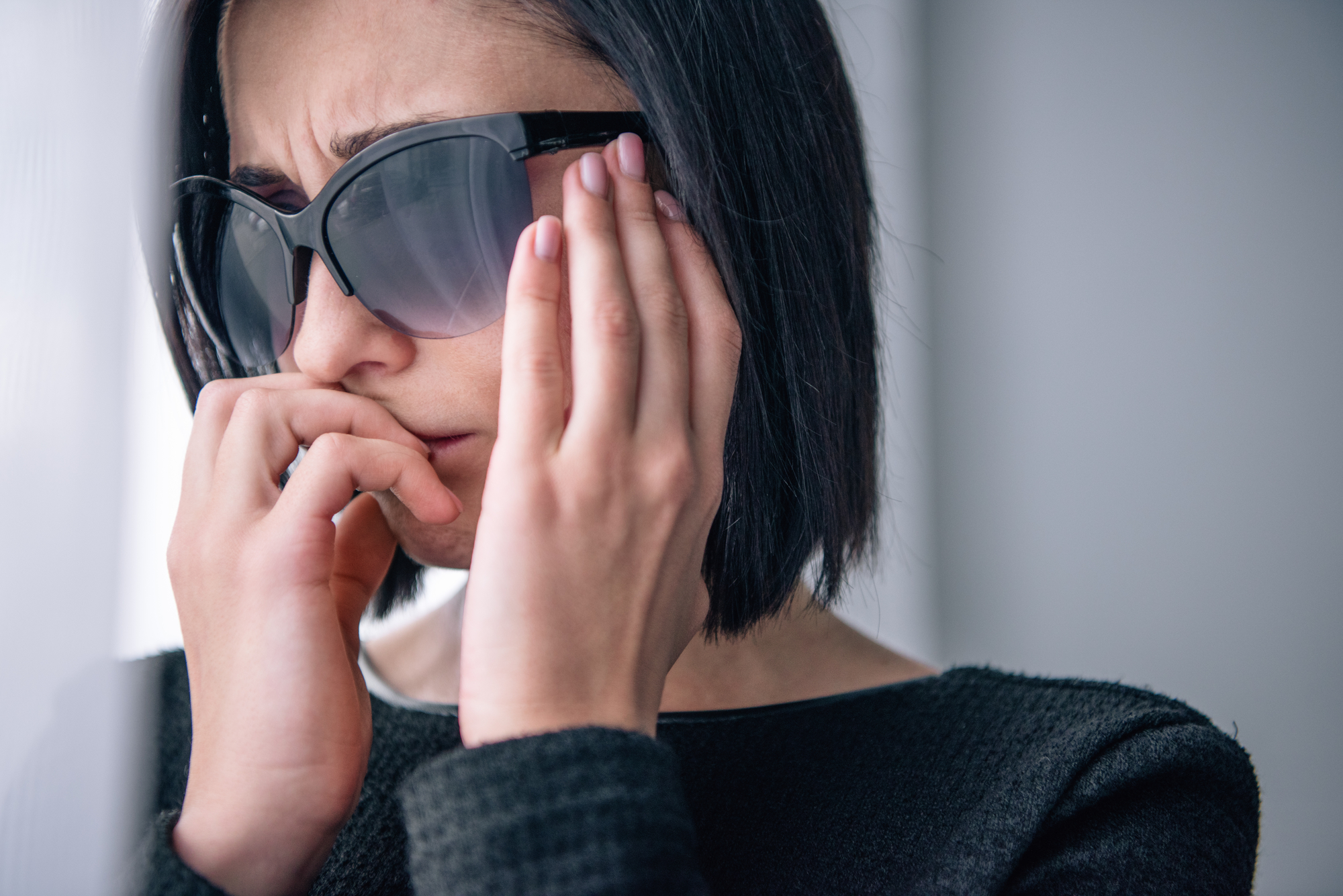 A person with short dark hair, wearing sunglasses and a dark top, holds their forehead and bites their nails with a worried expression. The background is blurry, focusing attention on the individual's anxious demeanor.