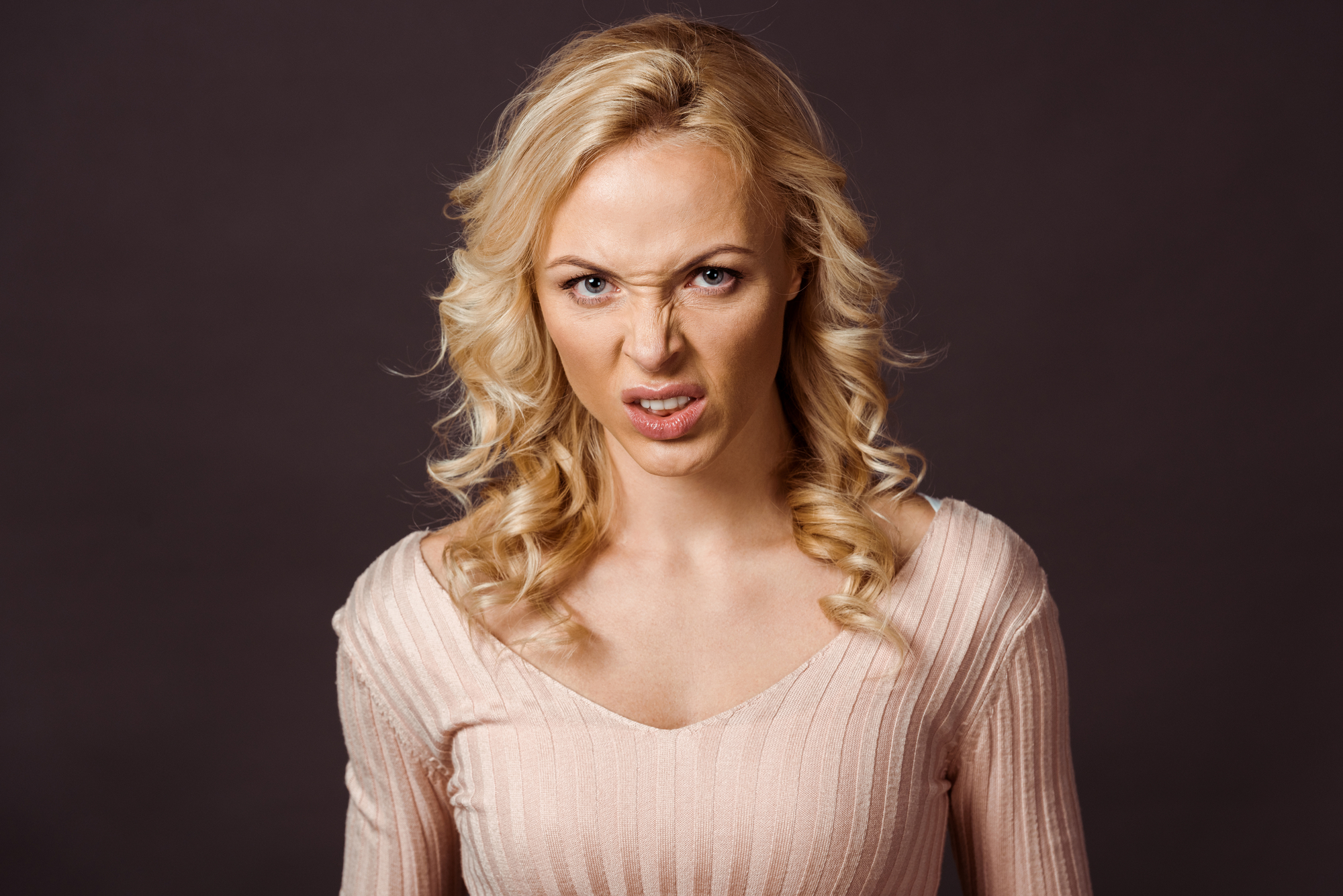 A person with long, wavy, blonde hair and a light pink top is standing against a dark background. They are making an angry, disgusted facial expression with their nose wrinkled and mouth slightly open.
