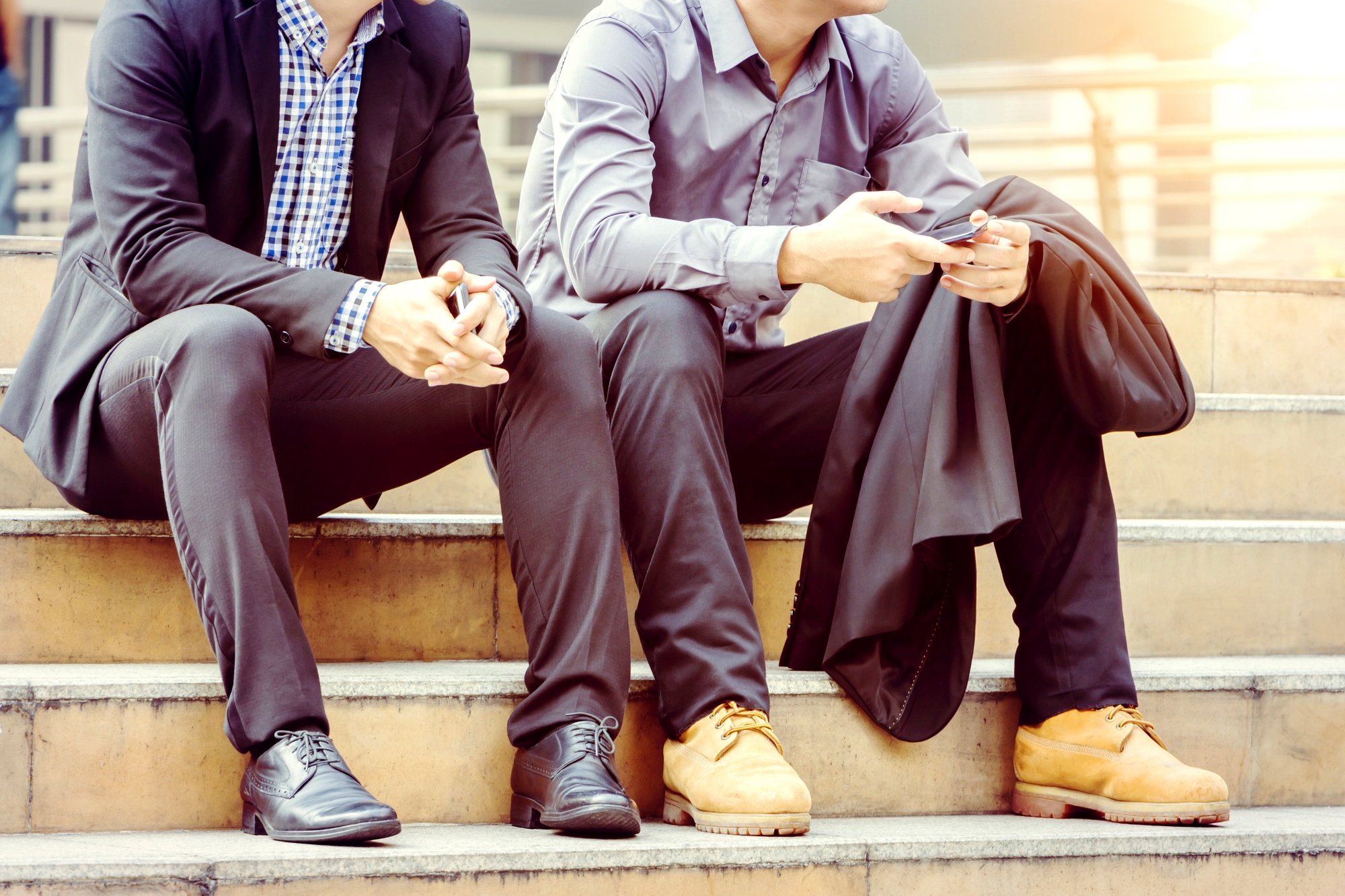 Two men sit on outdoor steps in semi-formal attire. One wears a black suit and striped blue shirt, while the other wears a gray shirt with black pants, holding a jacket and a phone. Sunlight illuminates the background, creating a warm atmosphere.