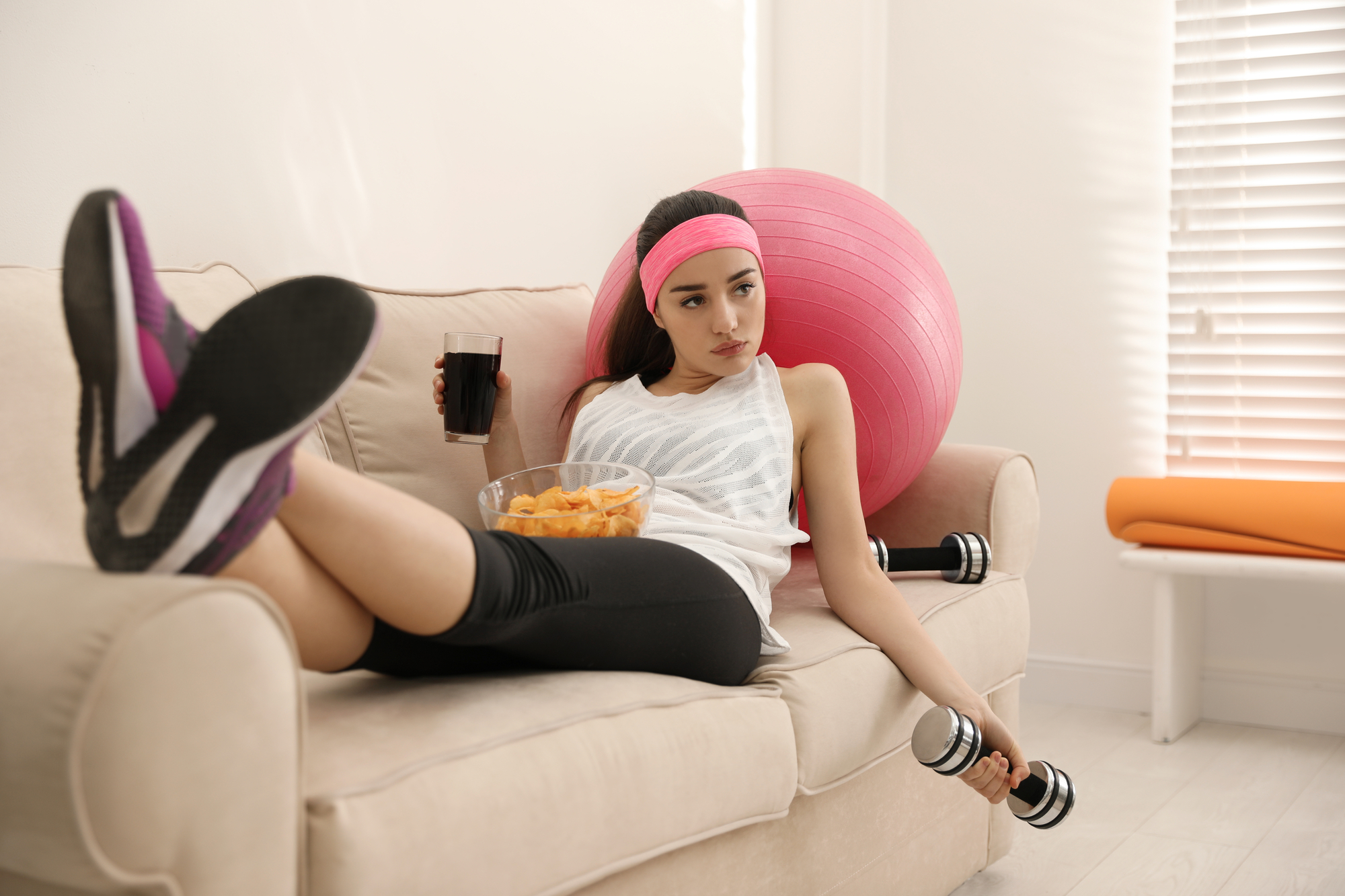 A woman in workout attire lounges on a beige sofa with a pink exercise ball. She holds a dumbbell in one hand and a bowl of snacks in the other, while a drink is placed nearby. She appears to be taking a break from exercising.